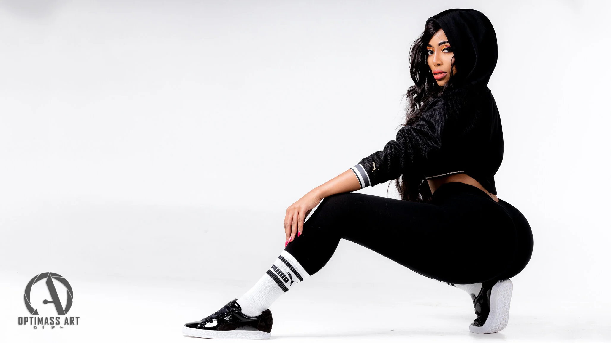 A woman is squatting with one knee on the ground, wearing black athletic clothing, white Puma socks, and black sneakers, with a black hooded jacket.