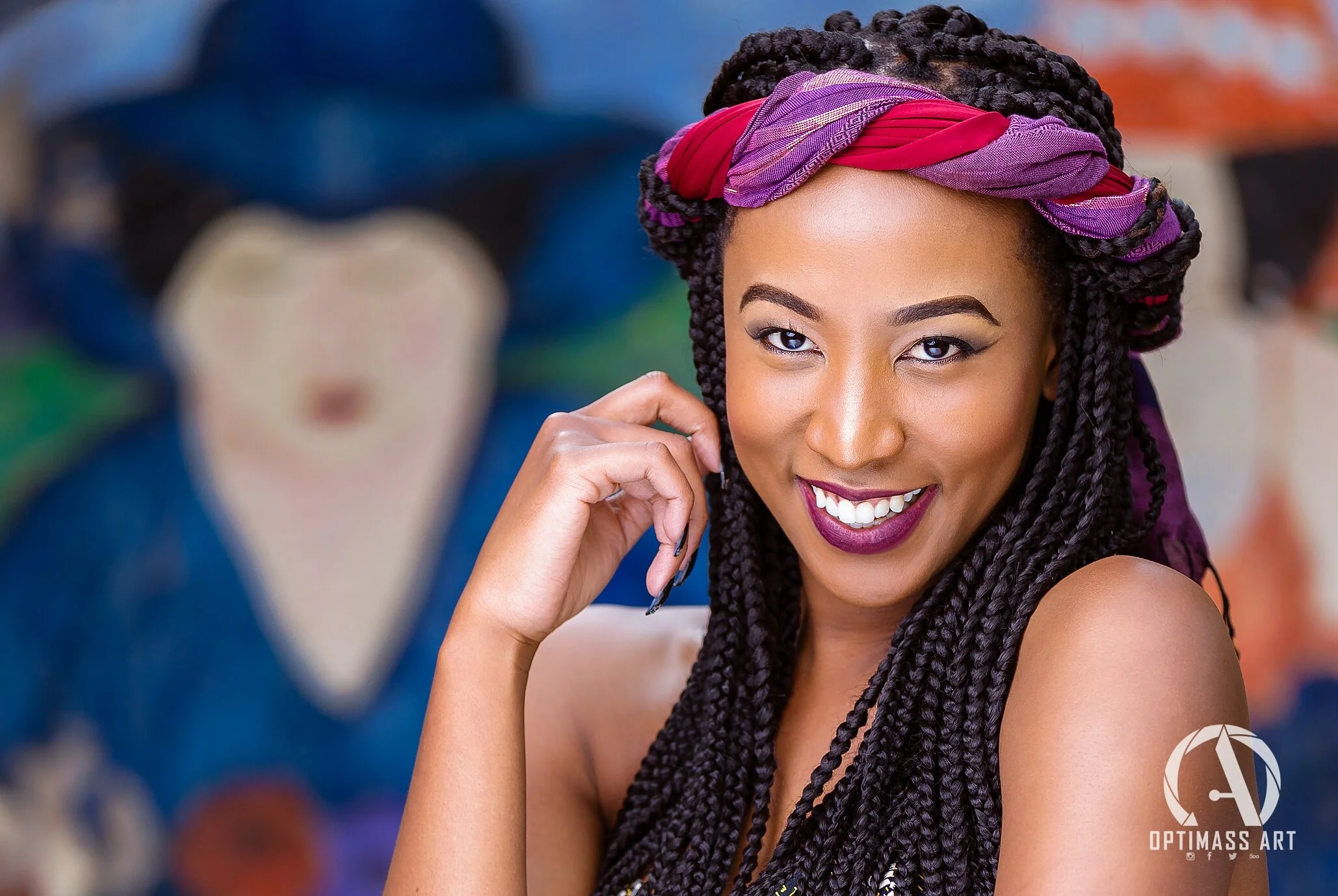 A woman with long braided hair wearing a purple and red headscarf, smiling with purple lipstick, resting her chin on her hand.