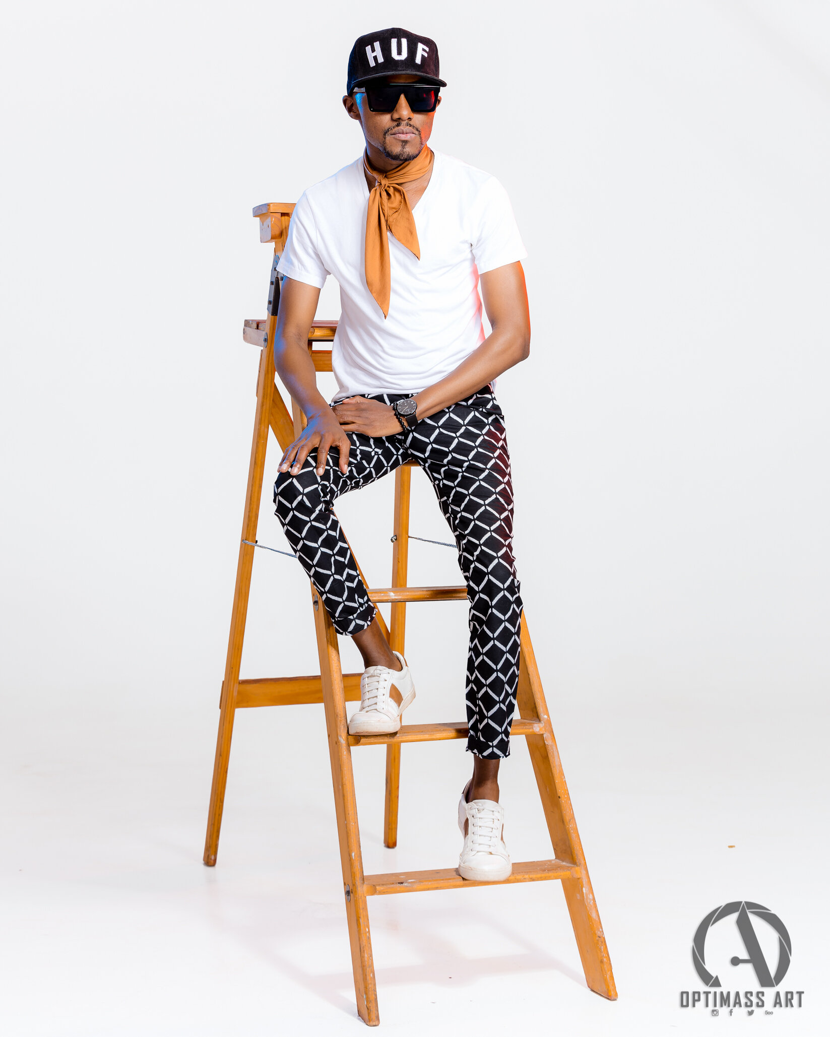 A man sitting on a ladder wearing sunglasses, a black cap with "HUF" written on it, a white t-shirt, patterned black and white pants, white sneakers, an orange scarf, and a wristwatch.