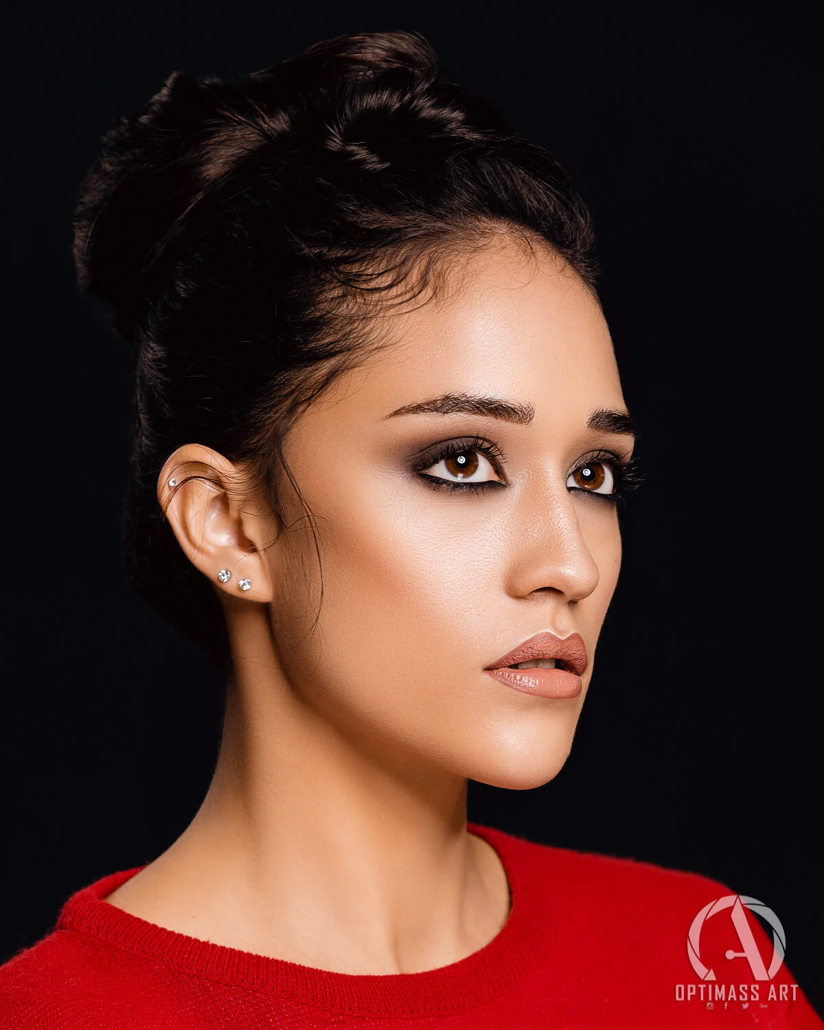 Close-up of a woman with dark hair styled upwards, wearing makeup with smoky eyeshadow and nude lipstick, earrings, and a red top, against a black background.