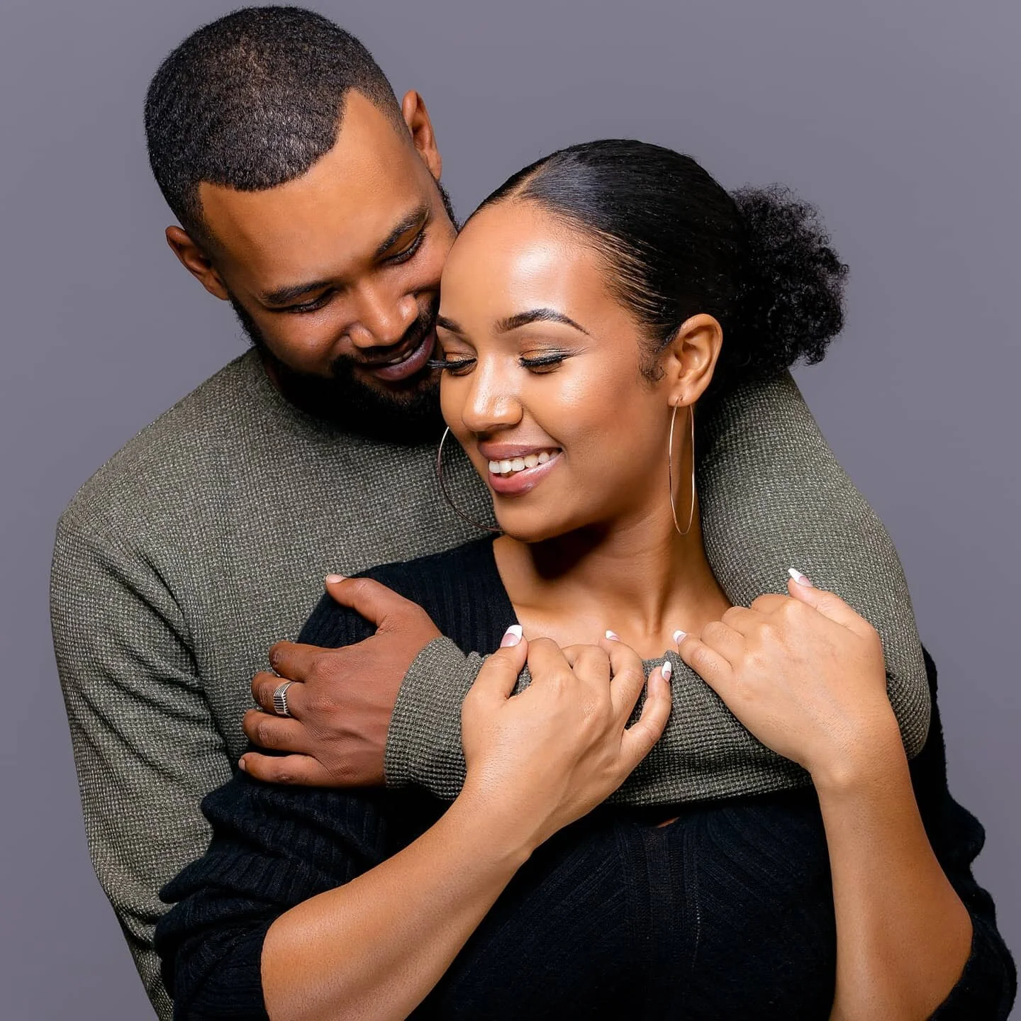 A couple hugging, smiling, and showing affection against a plain gray background.