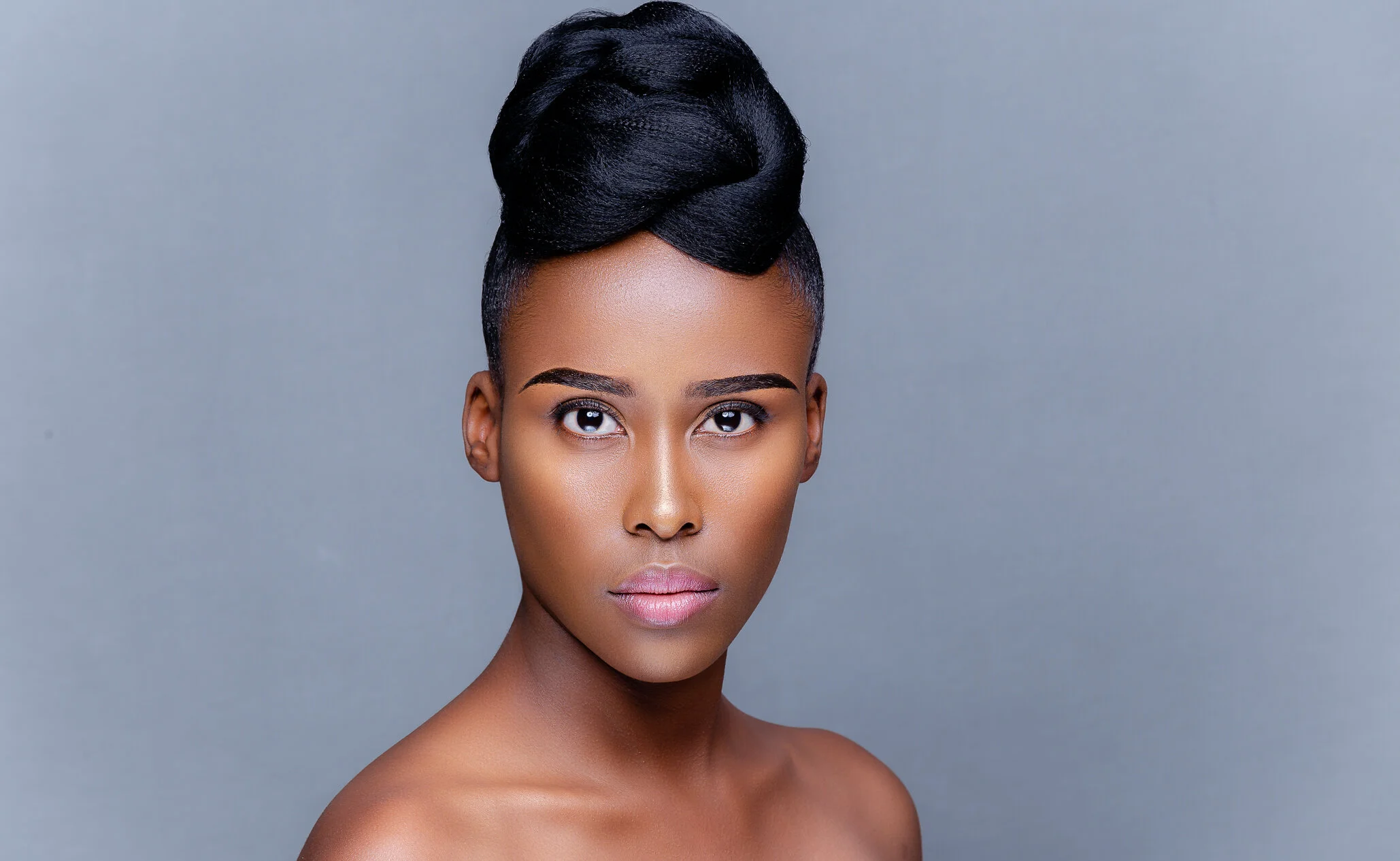 A portrait of a woman with dark skin, styled black hair in an elegant updo, and neutral makeup against a gray background.
