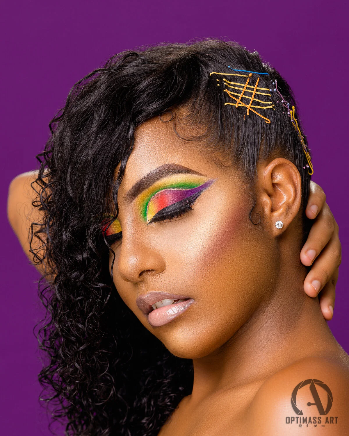 Close-up of a woman with rainbow-colored eye makeup, black curly hair with colorful hairpins, and a purple background.
