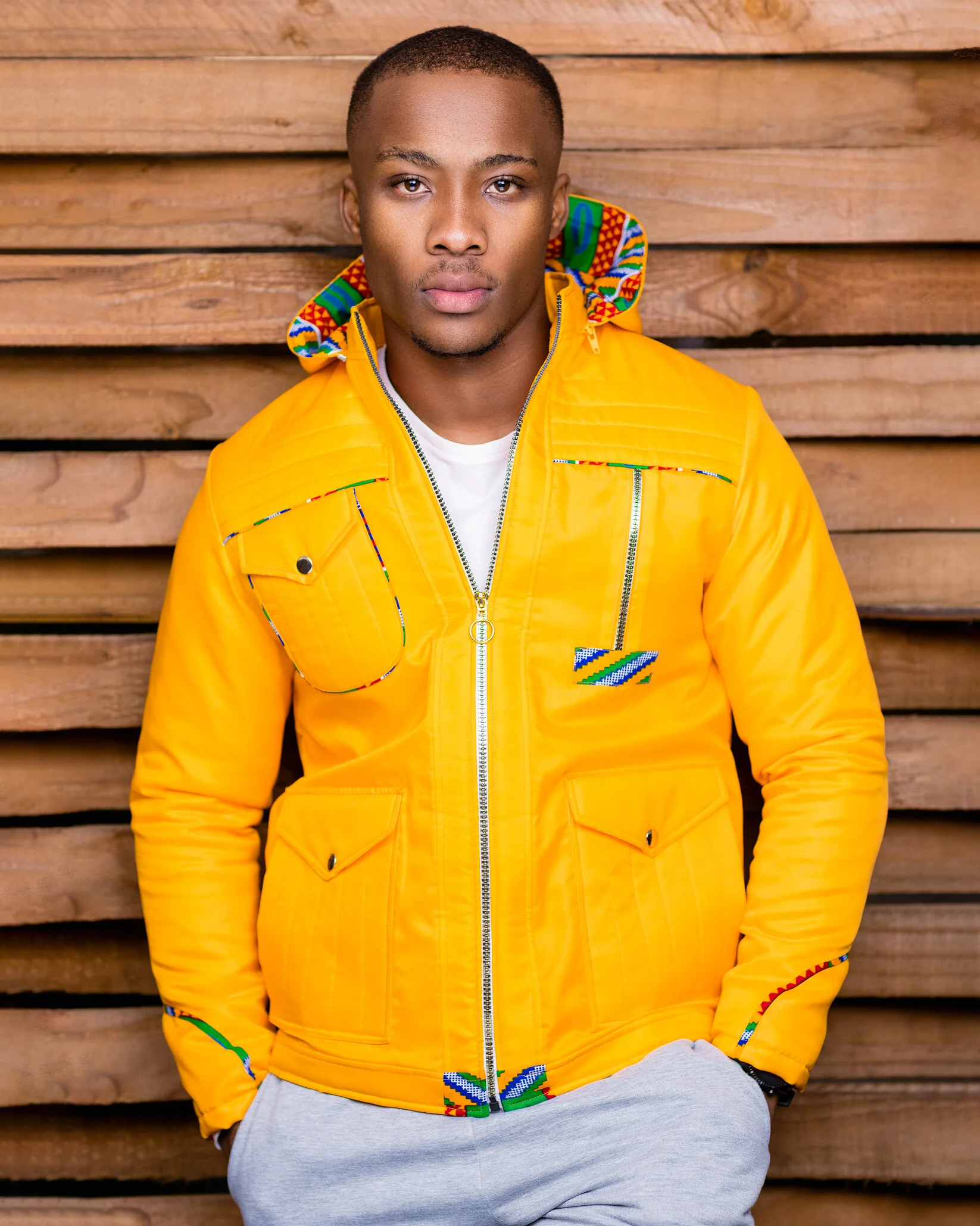 A young man with short hair wearing a bright yellow jacket with colorful beadwork accents, standing in front of a wooden fence.