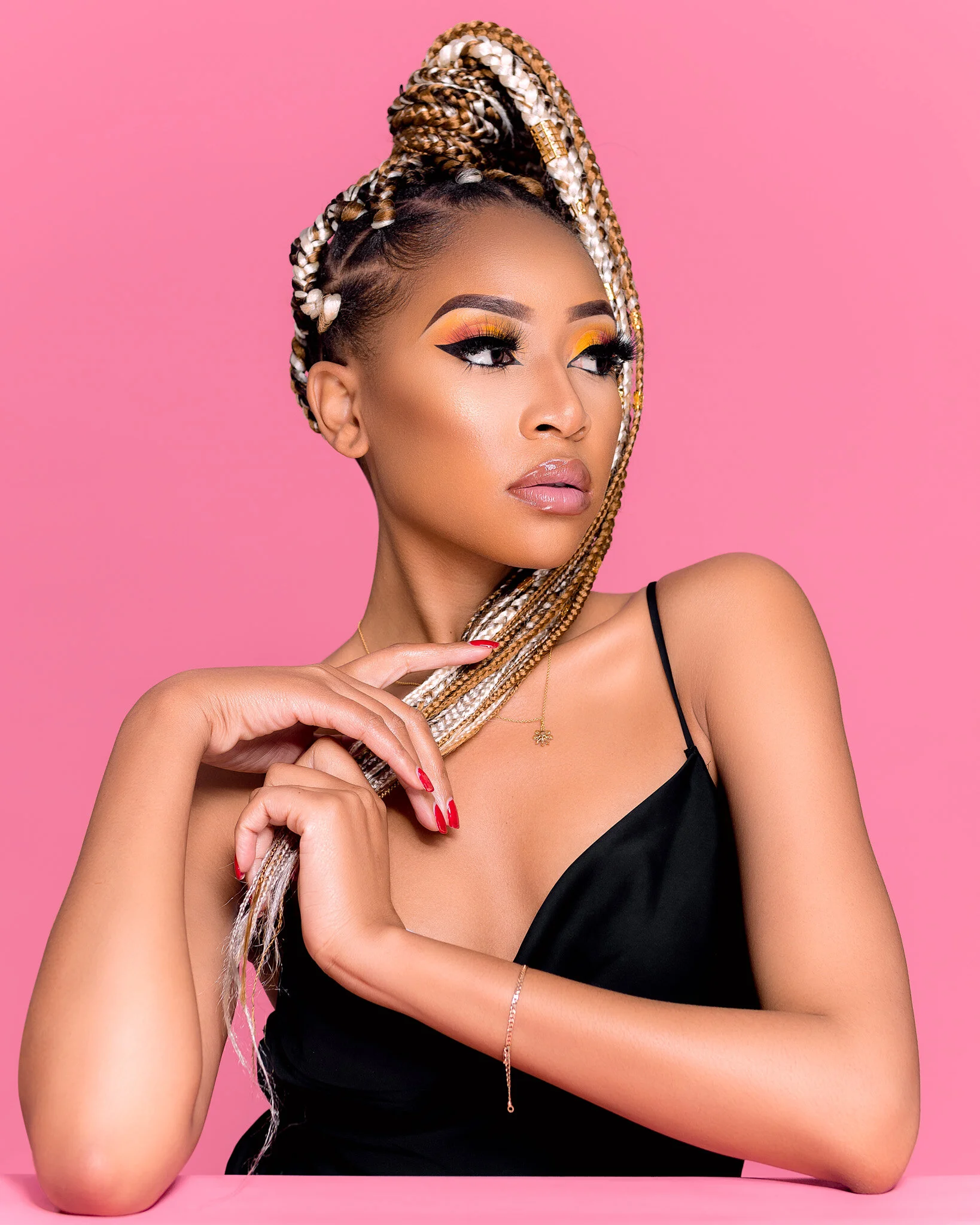 A woman with styled braids and makeup posed against a pink background, wearing a black spaghetti strap top, with her hands near her face and a gold necklace.