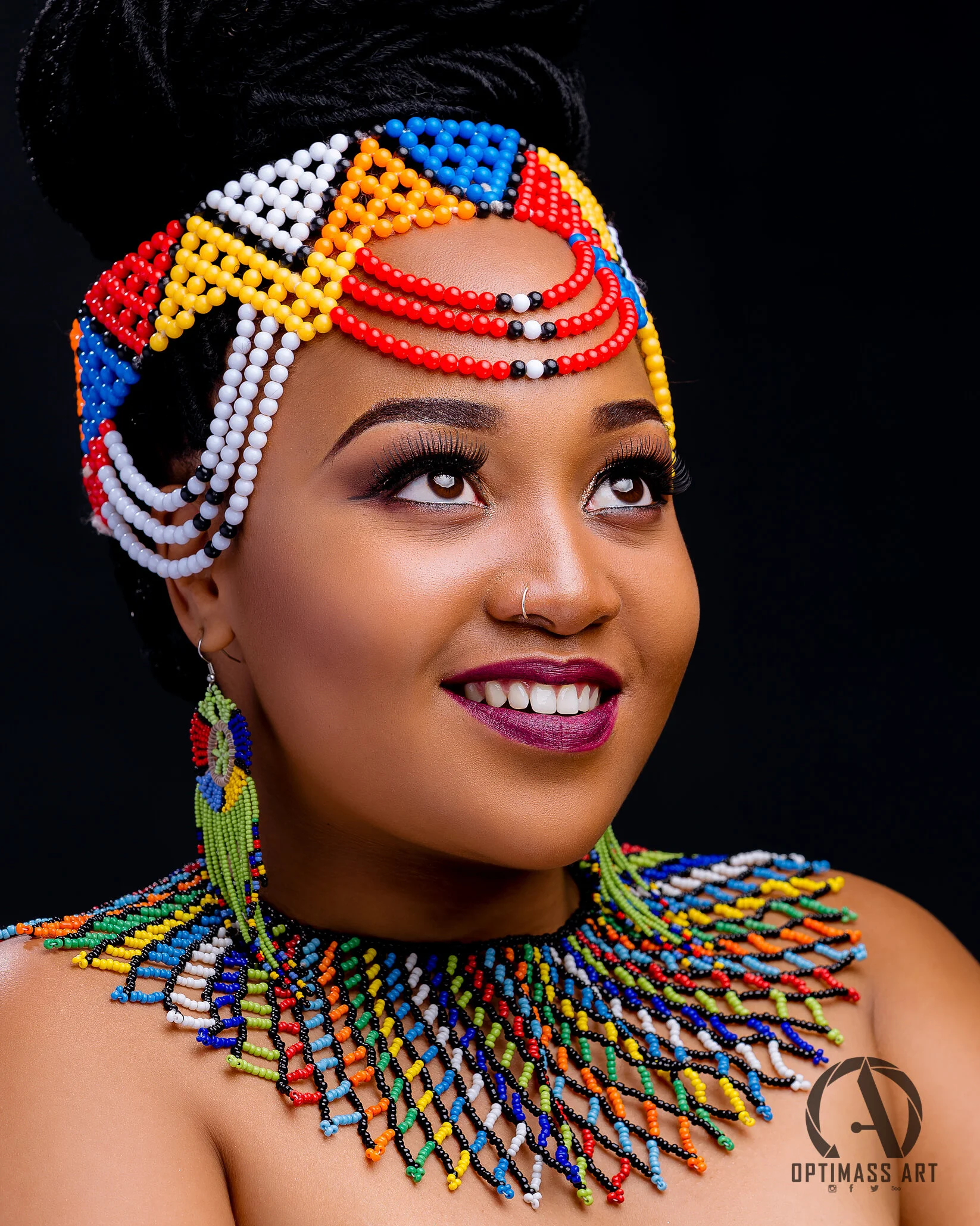 Portrait of a woman wearing colorful beaded jewelry and headwrap, smiling with makeup, against a black background.