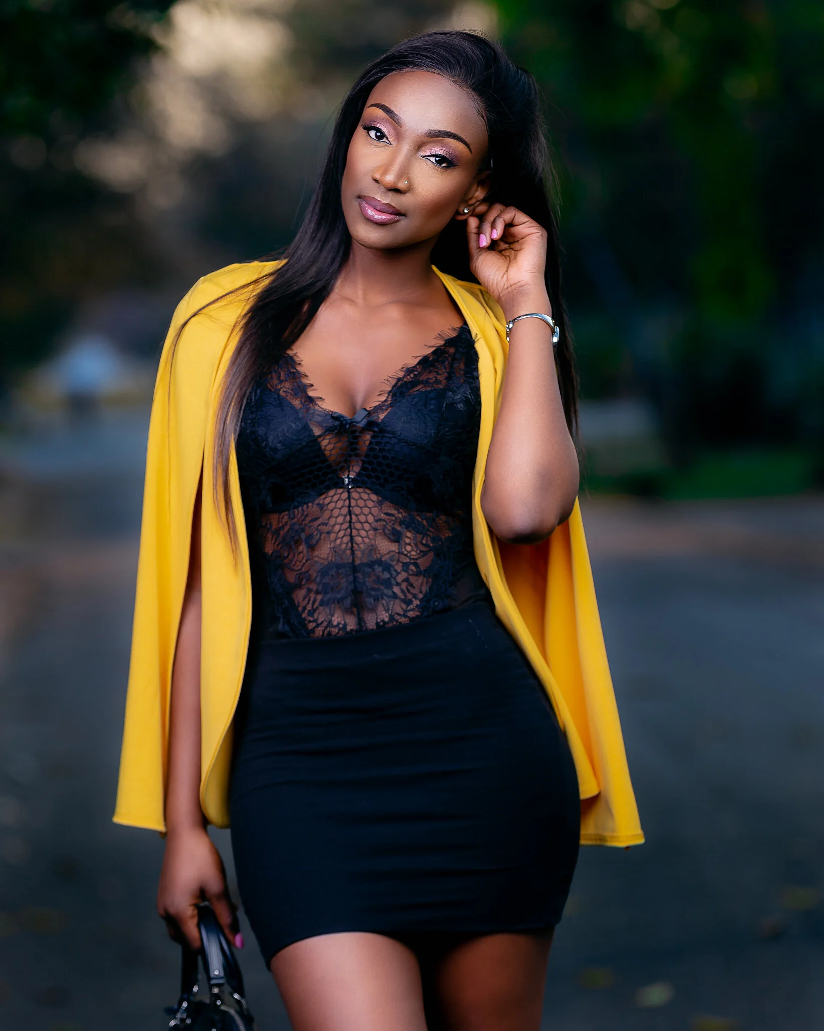 A woman with long black hair, dressed in a black lace top and a black skirt, wearing a yellow coat draped over her shoulders, standing outdoors with a blurred background of trees and dirt ground.