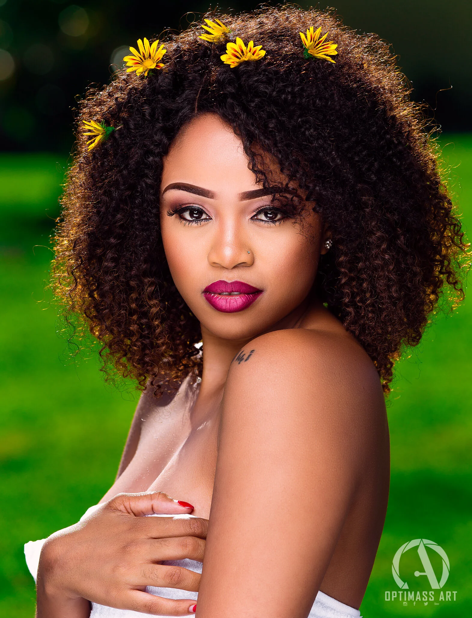 A woman with dark curly hair adorned with small yellow flowers, wearing makeup with bold lipstick, posing outdoors with a blurred green background.