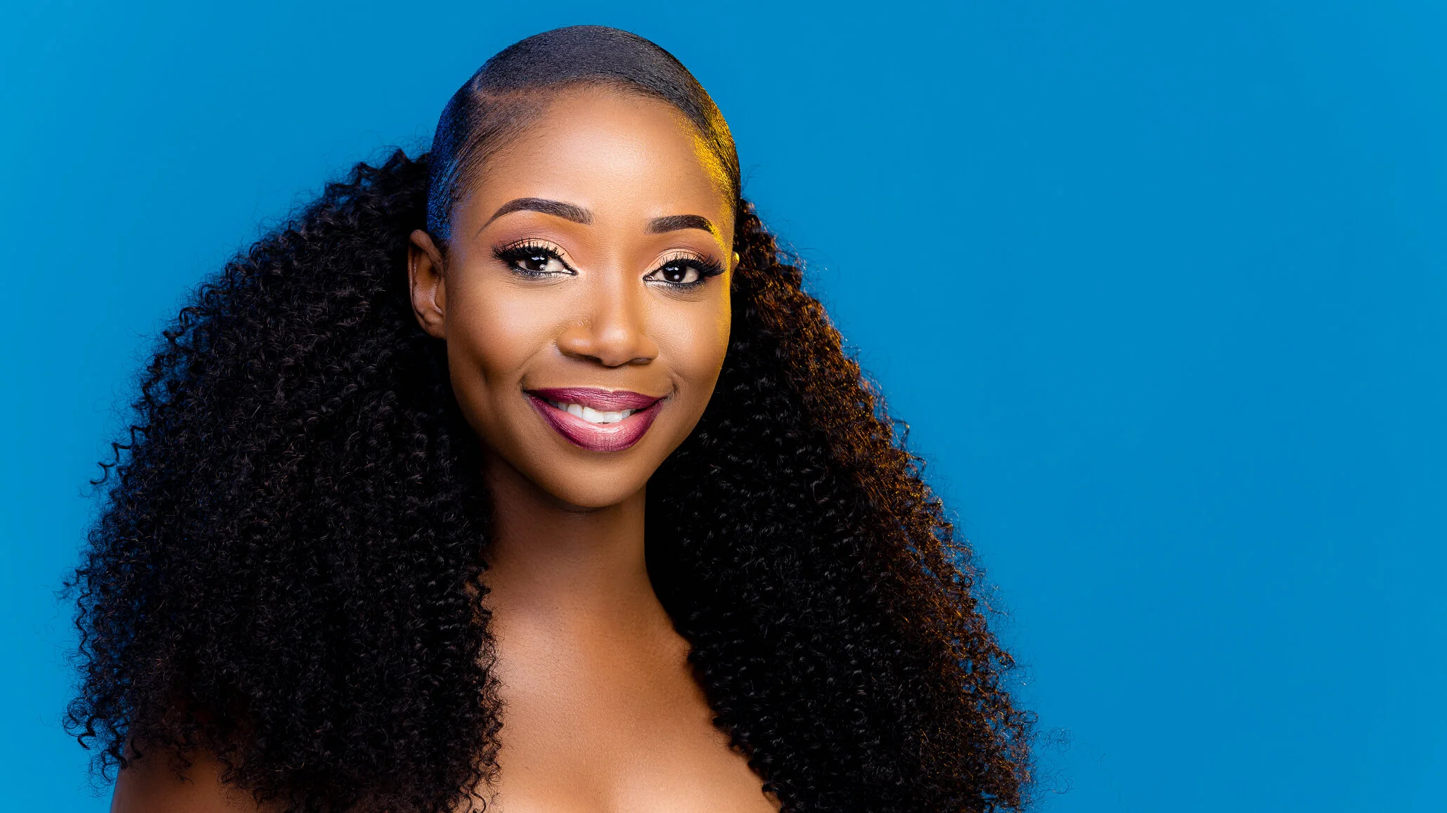 Portrait of a smiling woman with curly black hair, wearing makeup, set against a blue background.