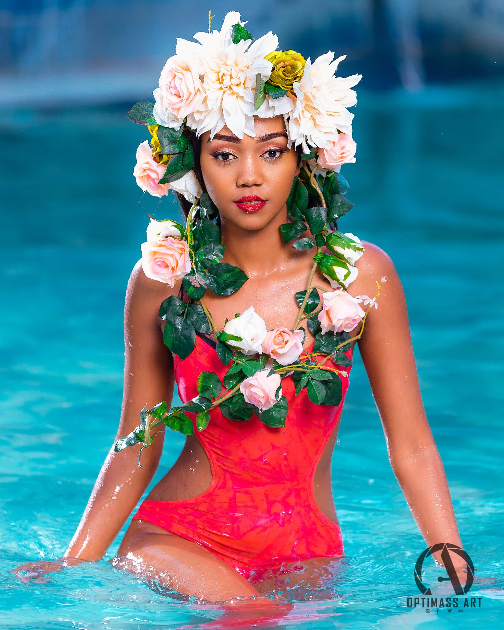 A woman in a red swimsuit standing in a pool, wearing a large floral headpiece and matching floral accessories around her neck and shoulders.