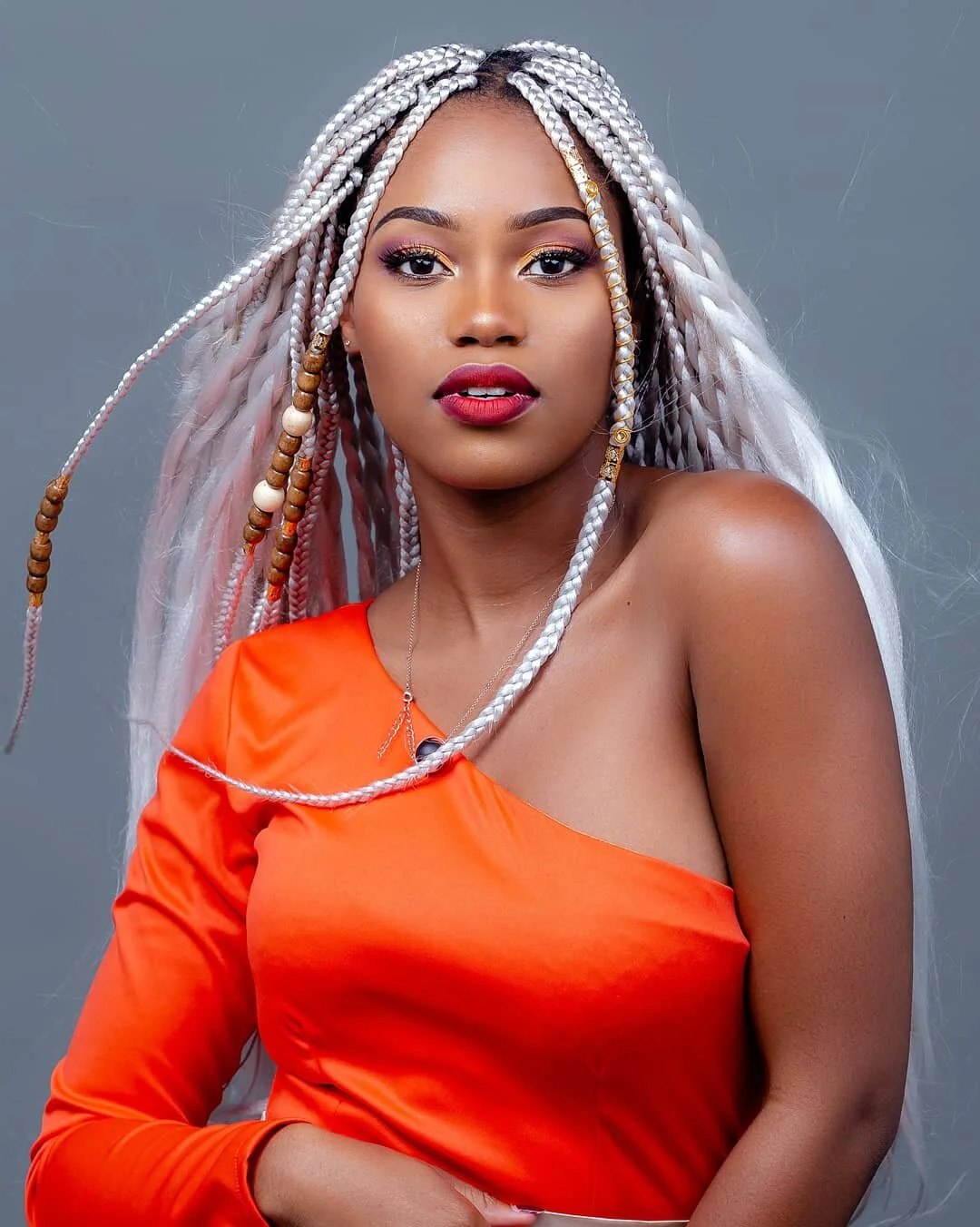 A woman with long braided silver and gold hair, wearing an orange one-shoulder top, red lipstick, and gold eye makeup, posing against a gray background.