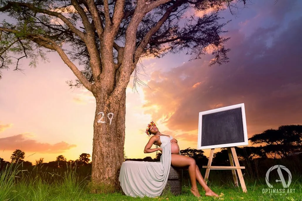 A pregnant woman with flowers in her hair sitting outdoors near a large tree with the number 29 painted on it. She is wearing a white dress and leaning back on a tire, with a black chalkboard on an easel nearby. The background features a colorful sun