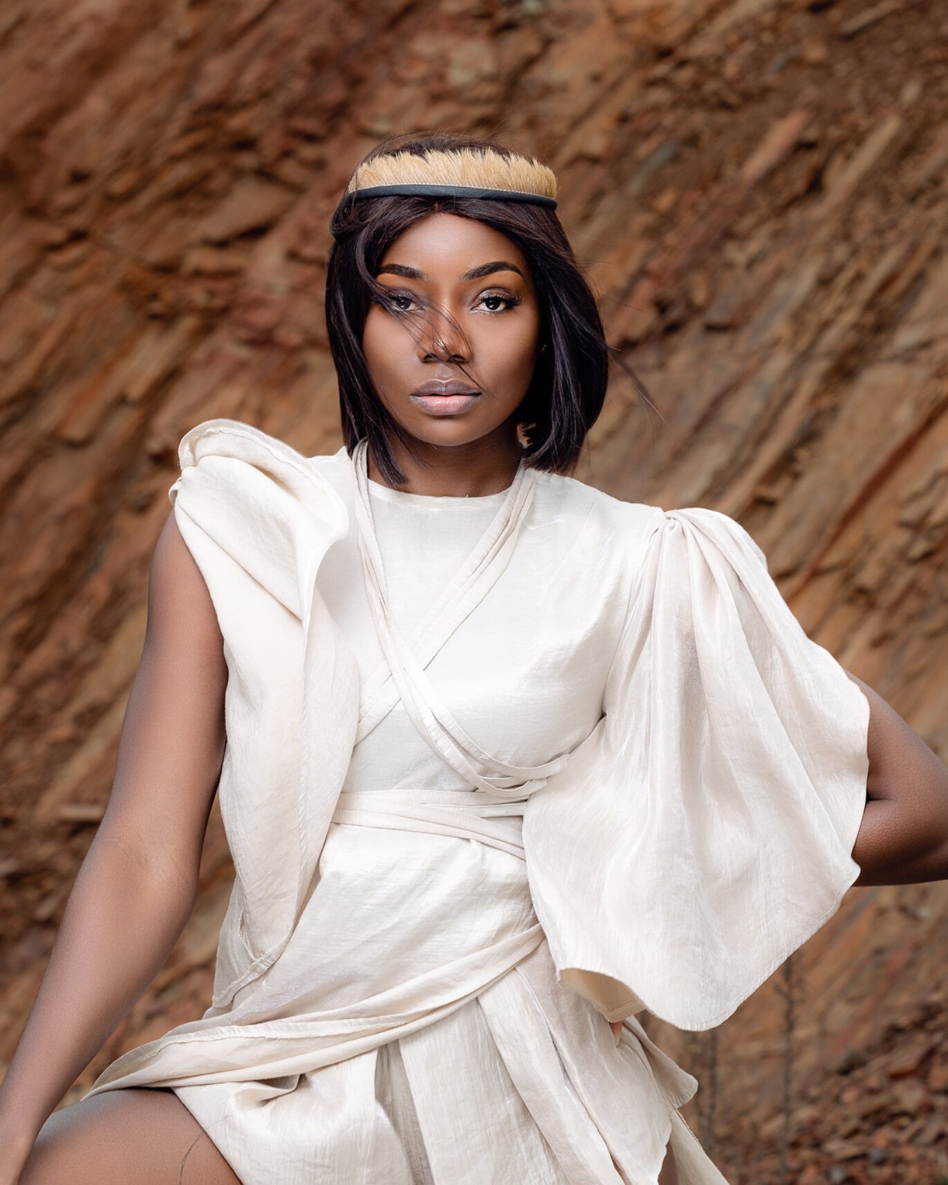 A woman with short black hair, wearing a beige headband and cream-colored dress with puffy sleeves, standing against a rocky background.