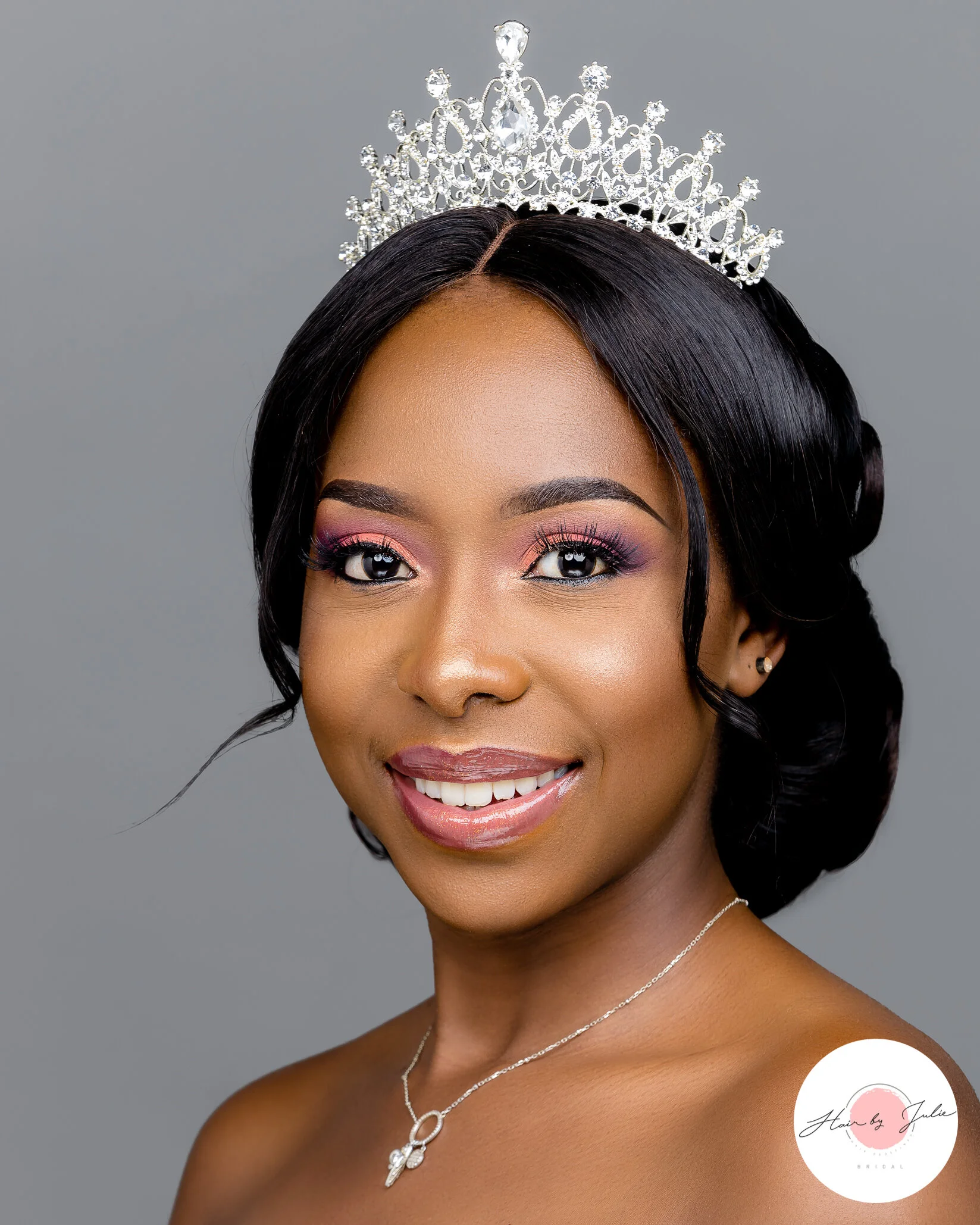 Portrait of a young African American woman wearing a tiara and jewelry, with styled black hair and makeup, smiling at the camera against a plain background.