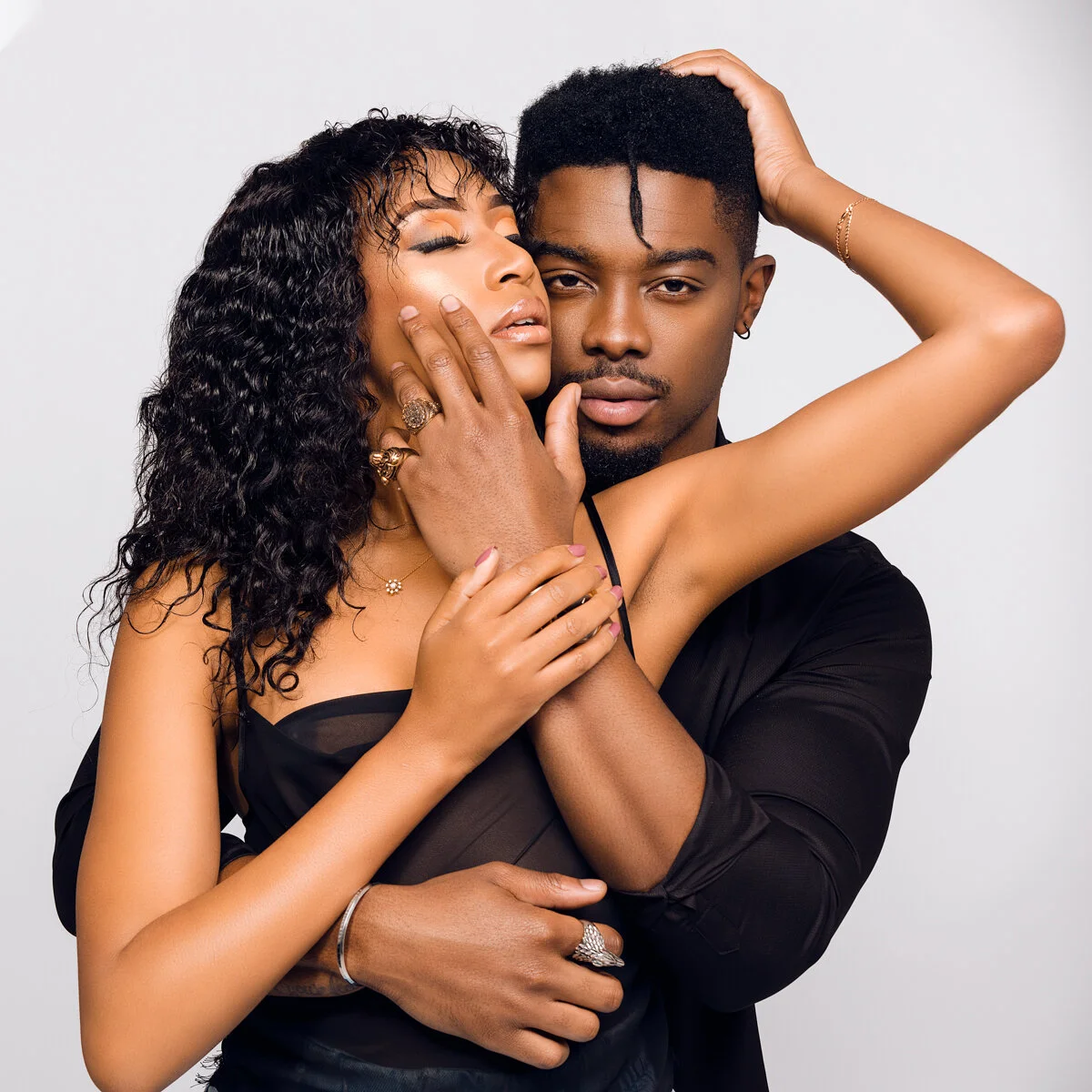 A woman with curly hair and a man with short hair embrace against a plain background, with the woman's arm around the man's neck and their faces close.