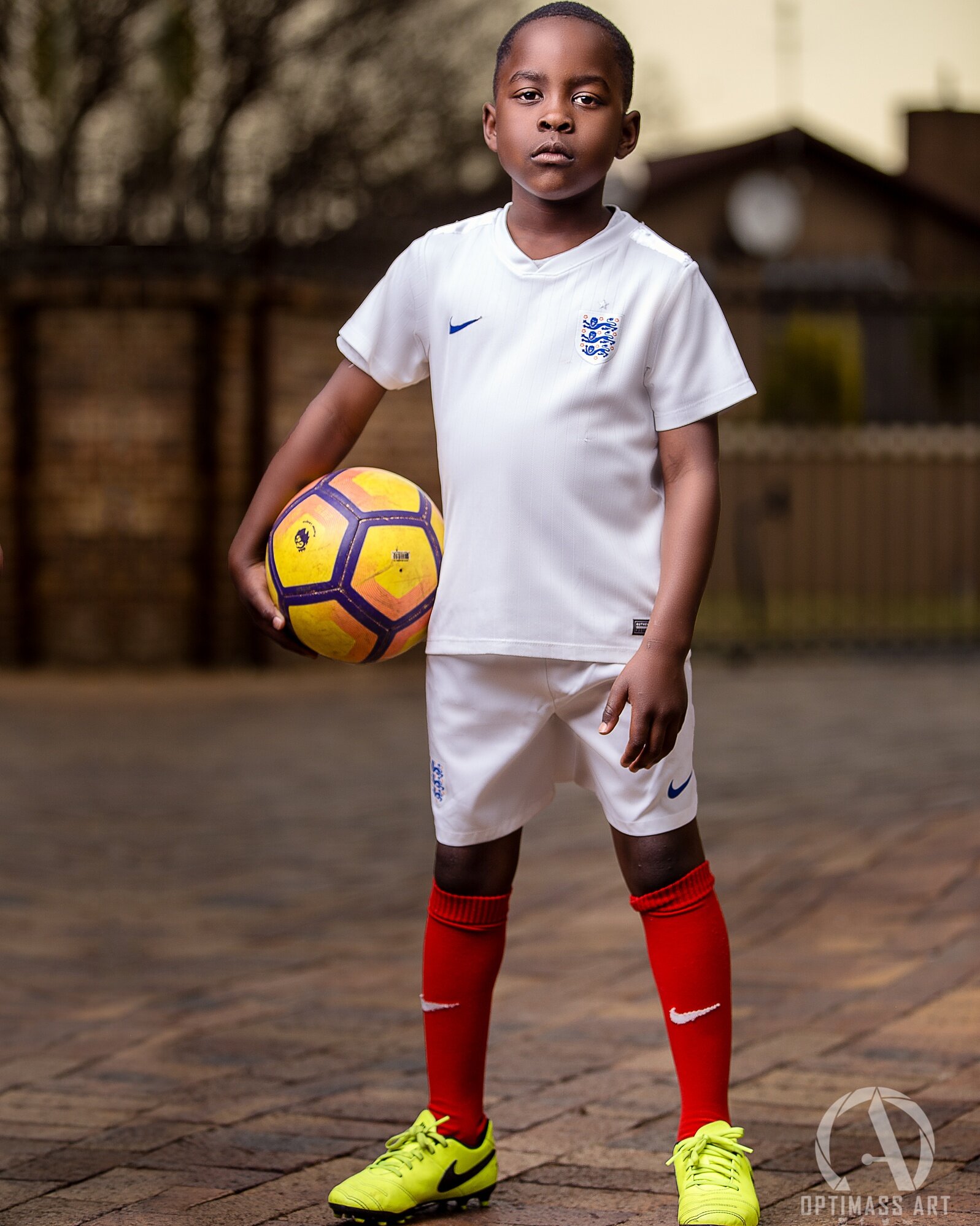 Young boy in white England soccer uniform holding a yellow and purple soccer ball, standing on a brick pavement outdoors.