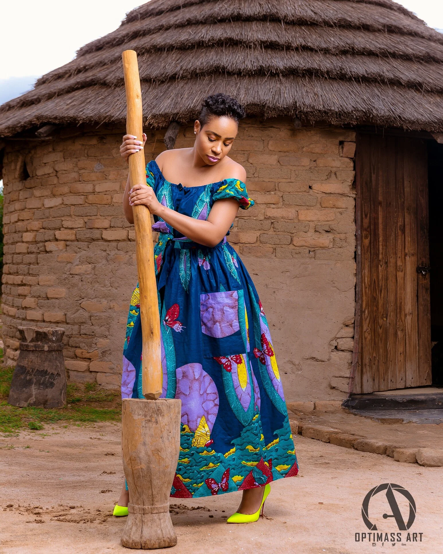 A woman in a colorful, butterfly and flower-patterned dress with yellow high heels, standing outside a round mud brick hut with a thatched roof, holding a large wooden mallet.