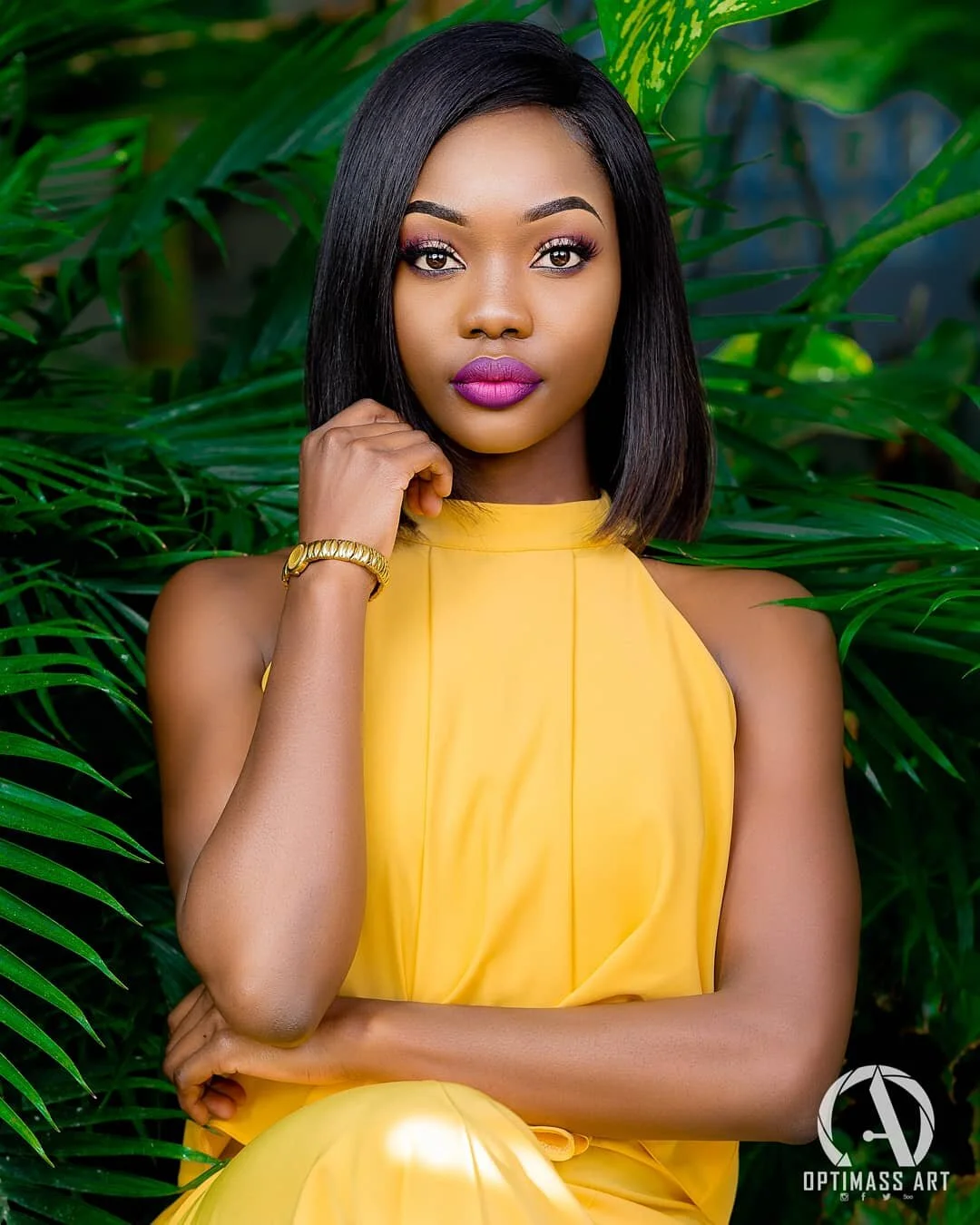 A young woman with straight black hair, wearing a yellow sleeveless dress, sitting among green tropical plants, with makeup including pink lipstick and eye makeup, and wearing a gold bracelet.