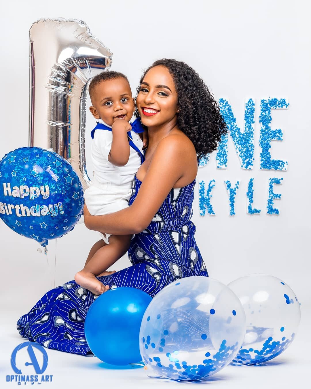 A woman holding a young boy during a birthday celebration with blue-themed balloons, a silver number 1 foil balloon, and a background with the words 'LOVE' and 'KYLE' made of blue pom-poms.