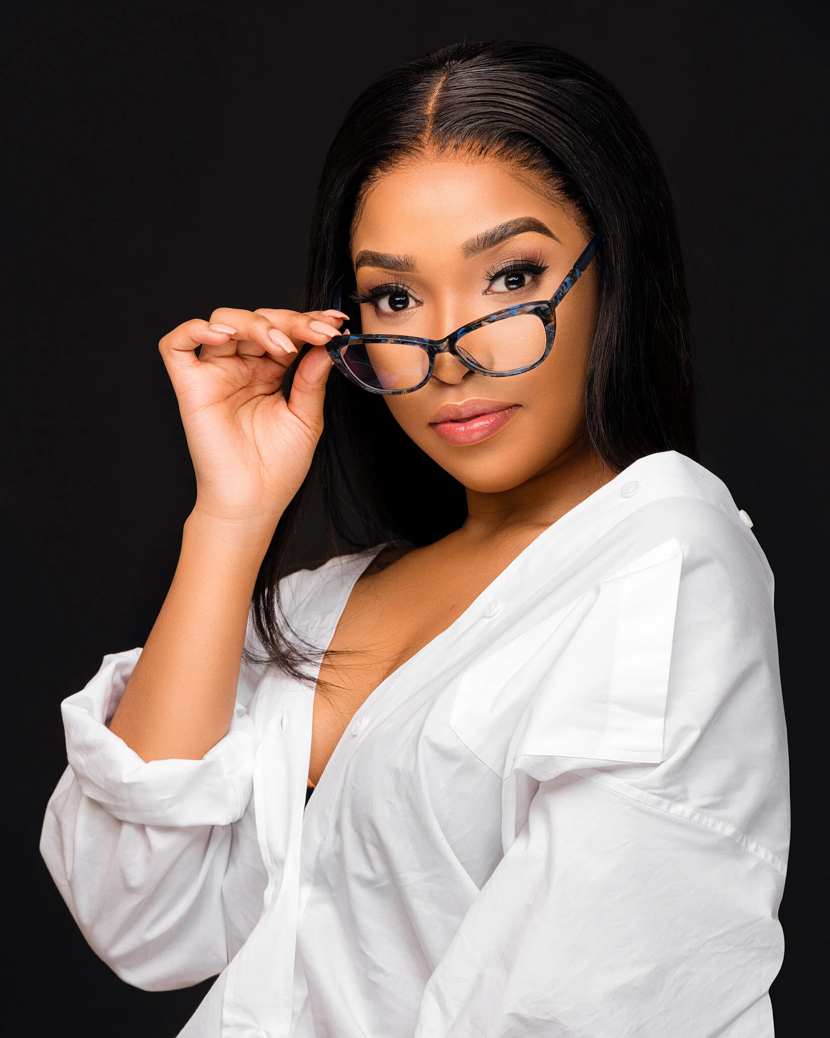 A woman with dark hair and makeup wearing a white shirt, holding glasses near her face against a black background.
