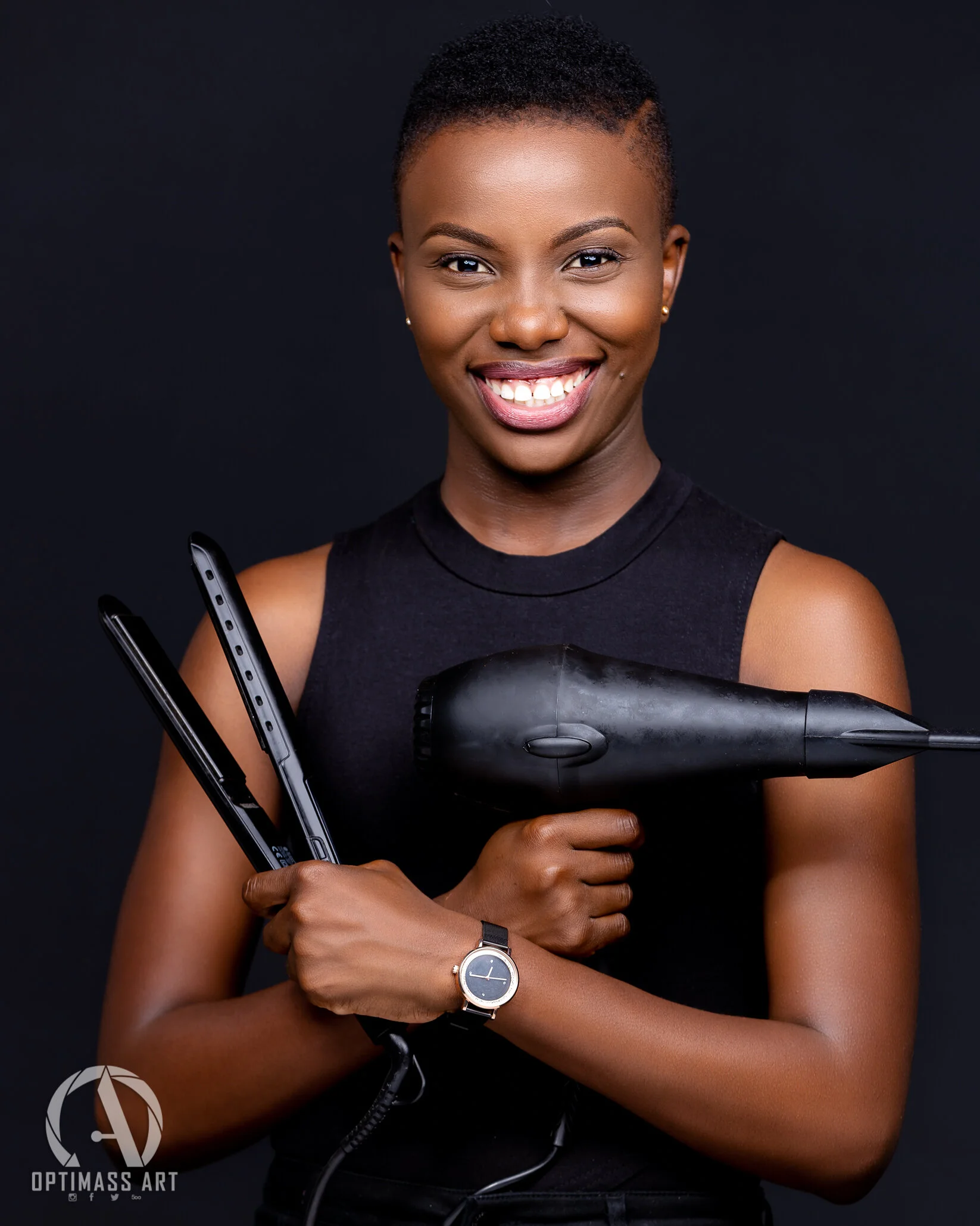 A smiling woman with short hair, wearing a black sleeveless top, holding a black hairdryer and a flat iron, against a black background.