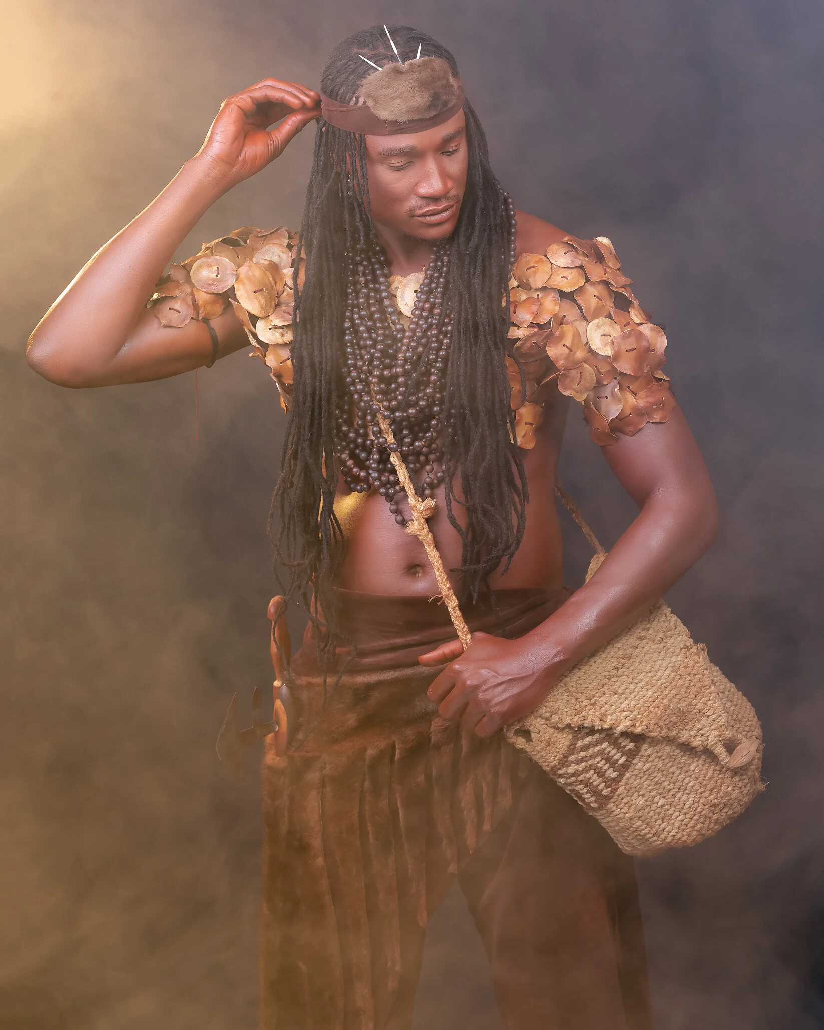 A person dressed in traditional attire with layered bead necklaces, flower-like shoulder decorations, and a woven bag, posing with a hand on their head against a smoky background.