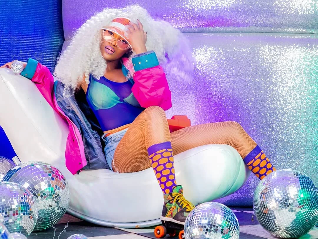 A person with curly white hair, pink glasses, and vibrant clothing sitting on a white chair, surrounded by disco balls, in front of a colorful glittery background, wearing roller skates.