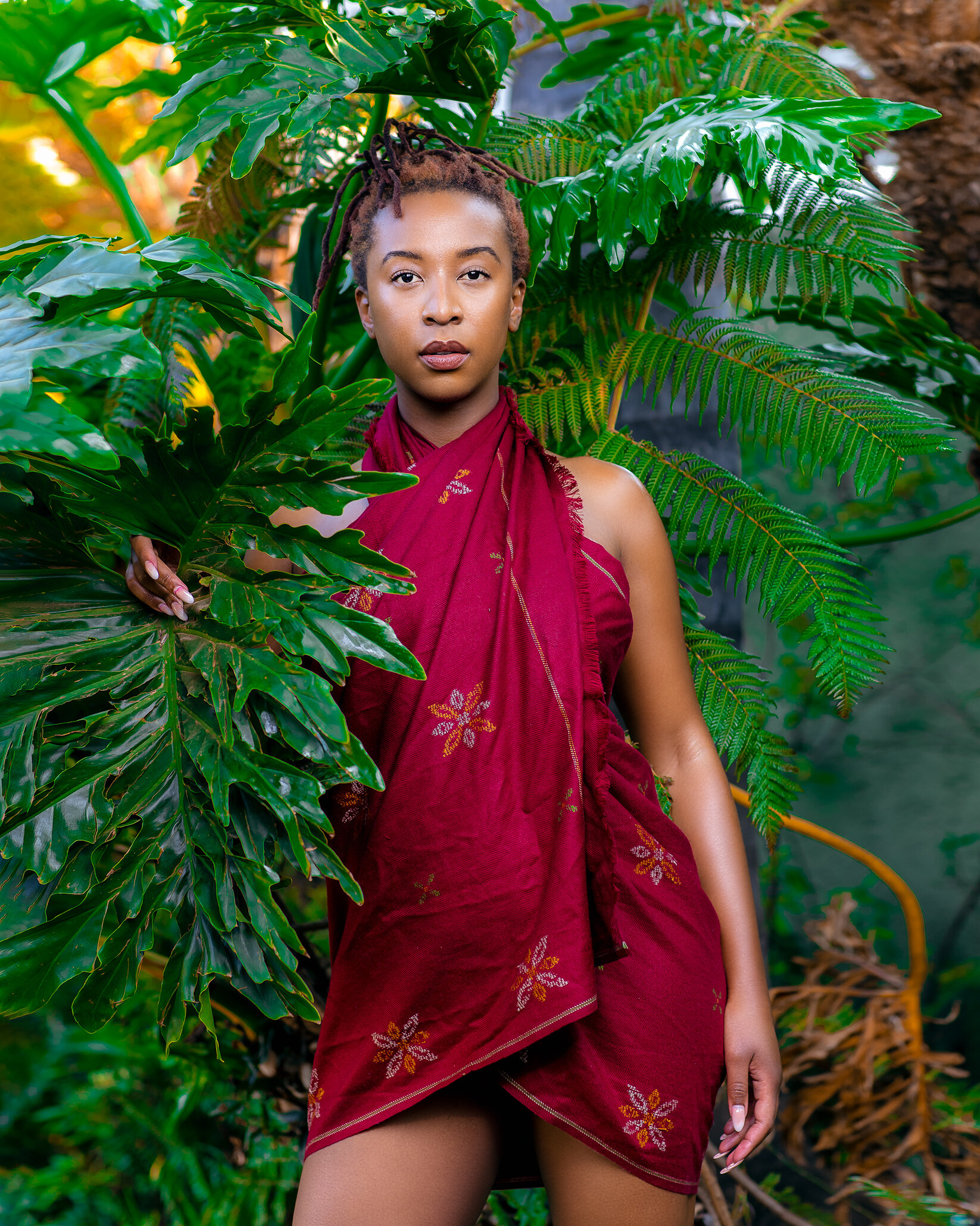 A woman with short dreadlocks and brown skin posing amid lush green tropical plants, wearing a red sarong with embroidered flowers.