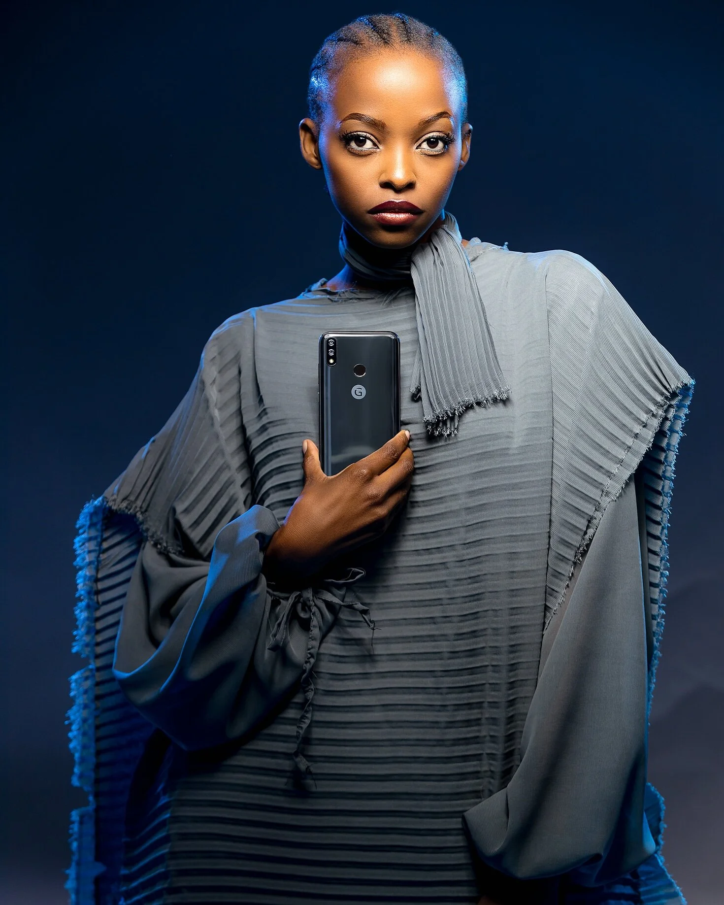 A woman with braided hair and dark lipstick holding a smartphone in front of her chest, wearing a pleated gray outfit with a scarf, against a dark background.