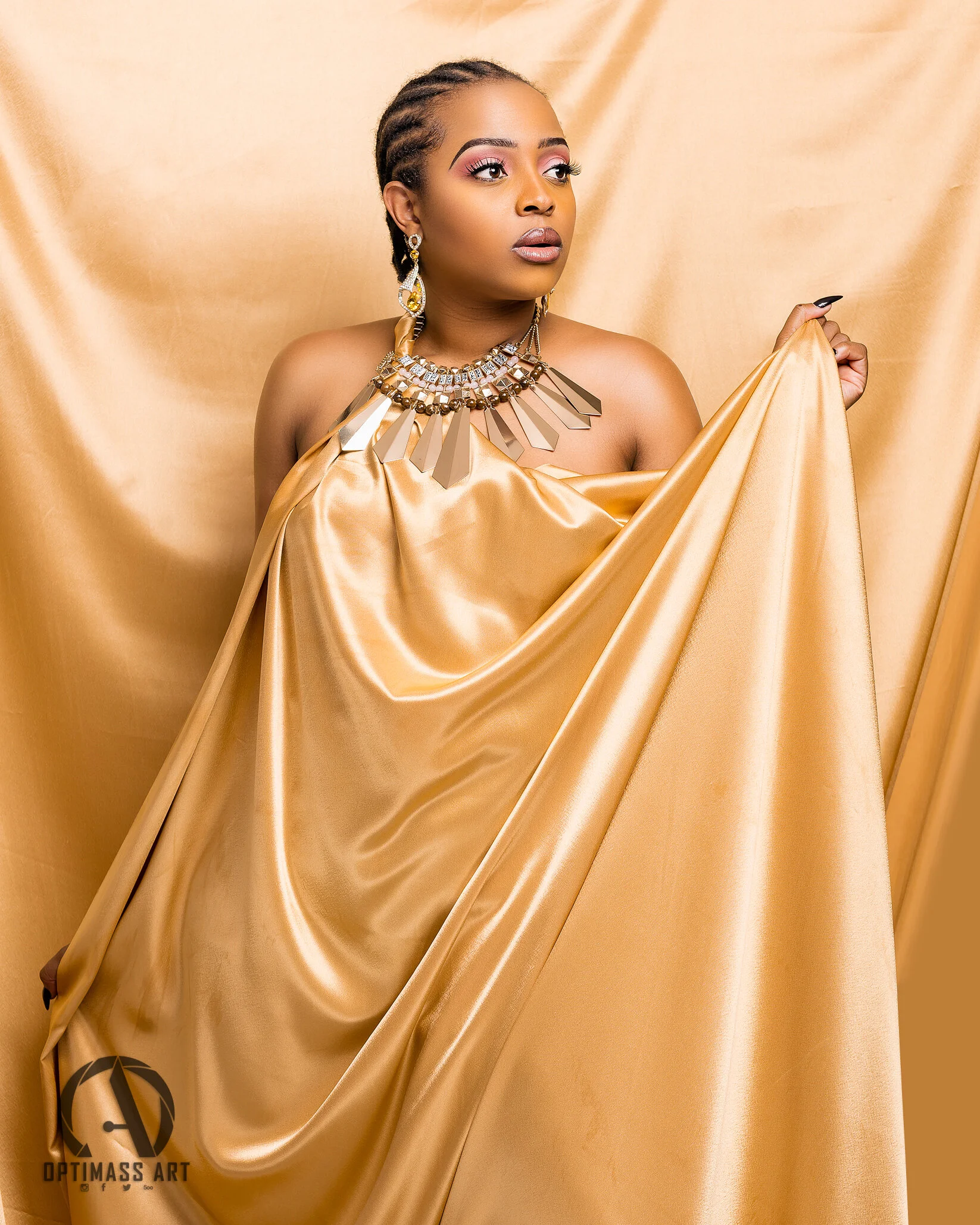 A woman with braided hair and jewelry stands against a satin gold backdrop, holding a matching gold satin fabric.