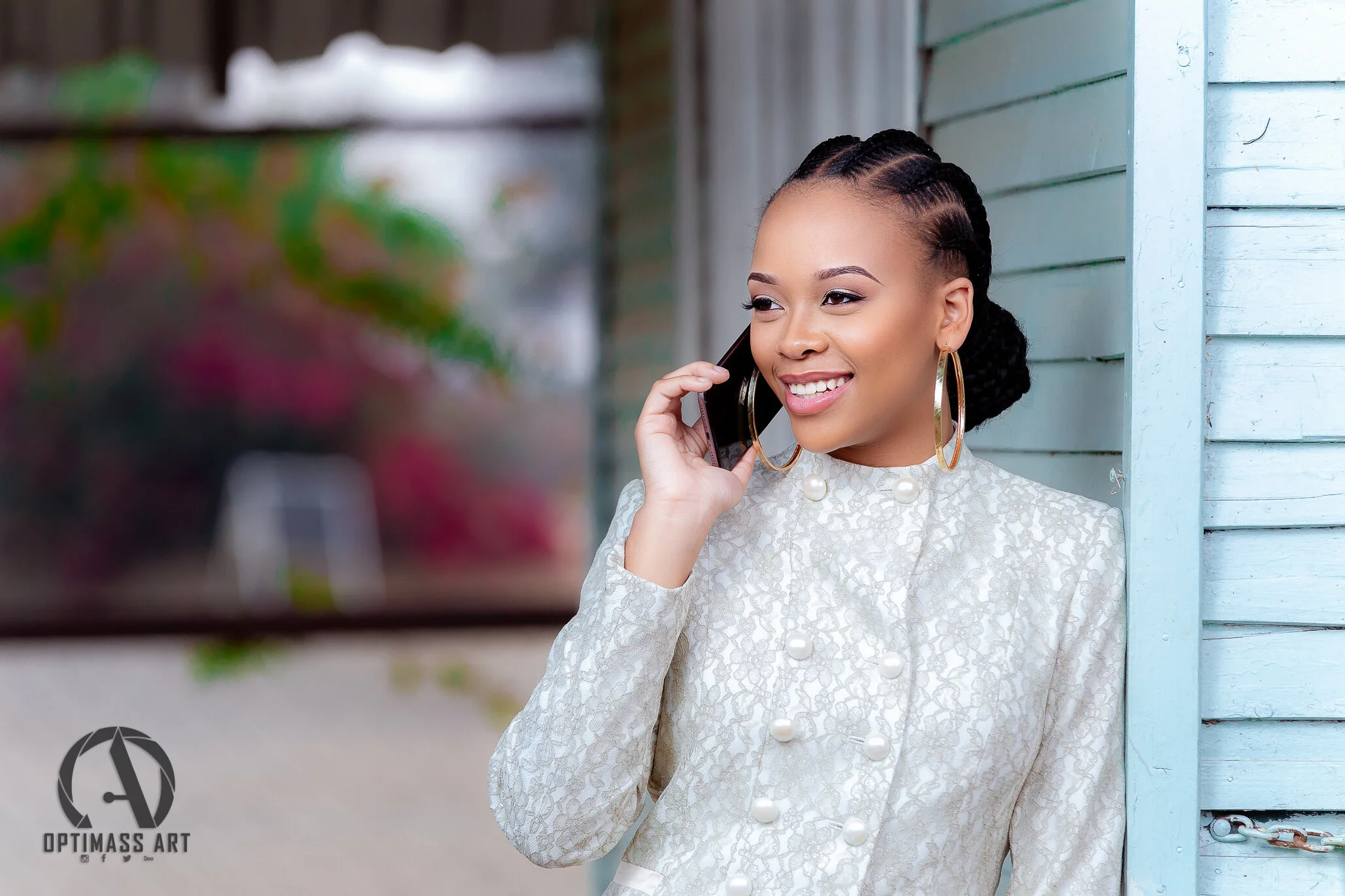 Young woman with braided hair, wearing a white lace jacket with pearl buttons and large hoop earrings, talking on a cellphone outdoors near a light blue wooden wall.