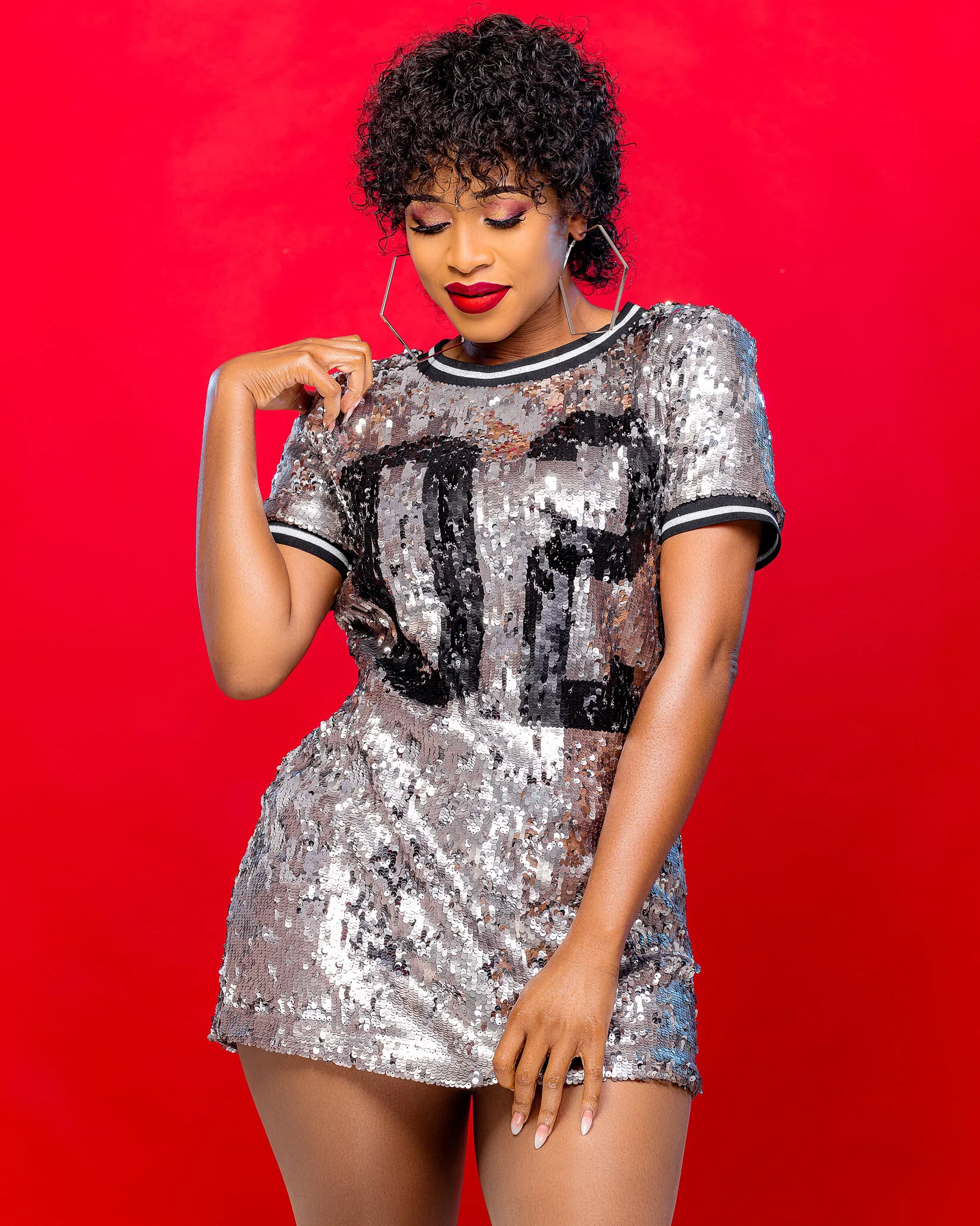 A woman wearing a silver sequined dress with a black and white collar, and hoop earrings, standing against a red background. She is looking down with her eyes closed and has short curly hair, red lipstick, and makeup.