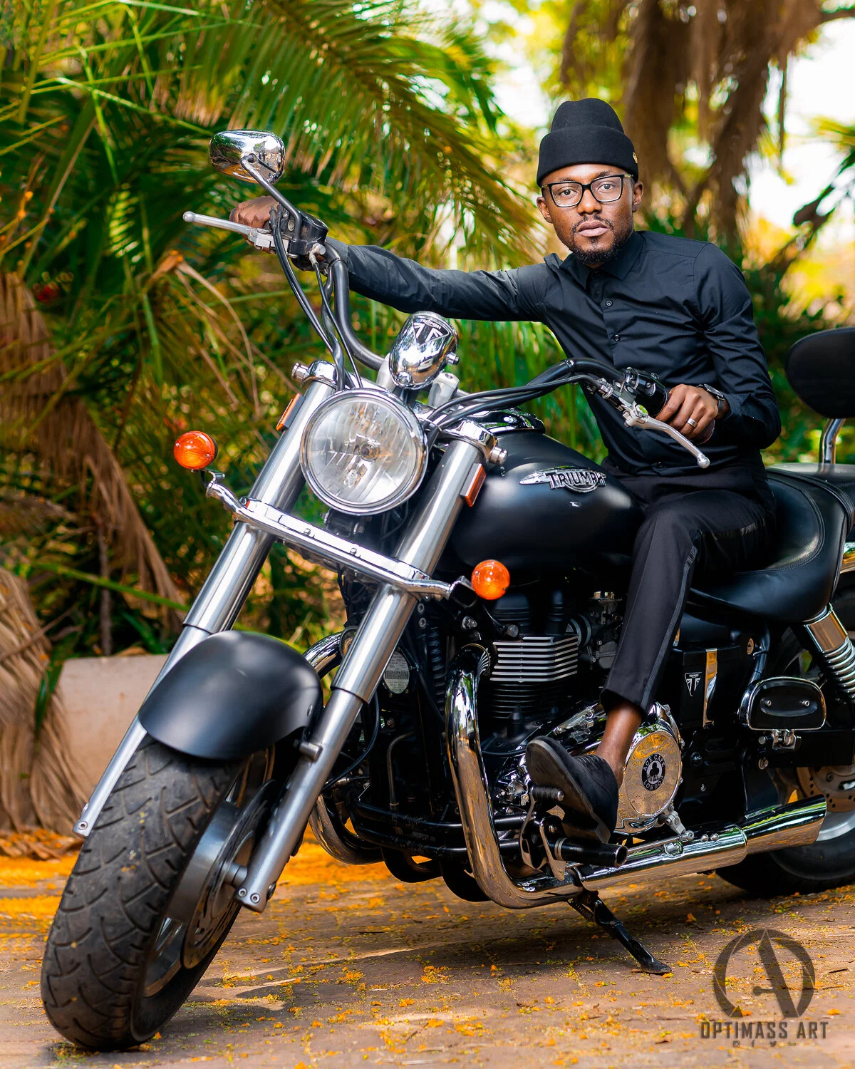 A man dressed in black sitting on a Triumph motorcycle outdoors surrounded by green foliage.