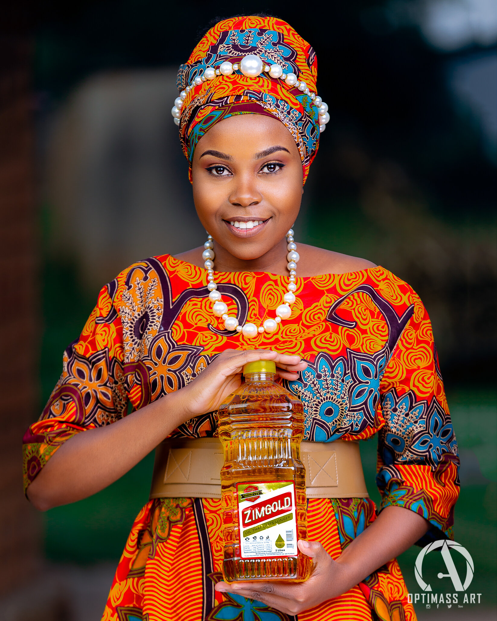 A woman wearing traditional African attire, including a vibrant headwrap and matching dress, holds a large bottle of Zimgold cooking oil and smiles at the camera.