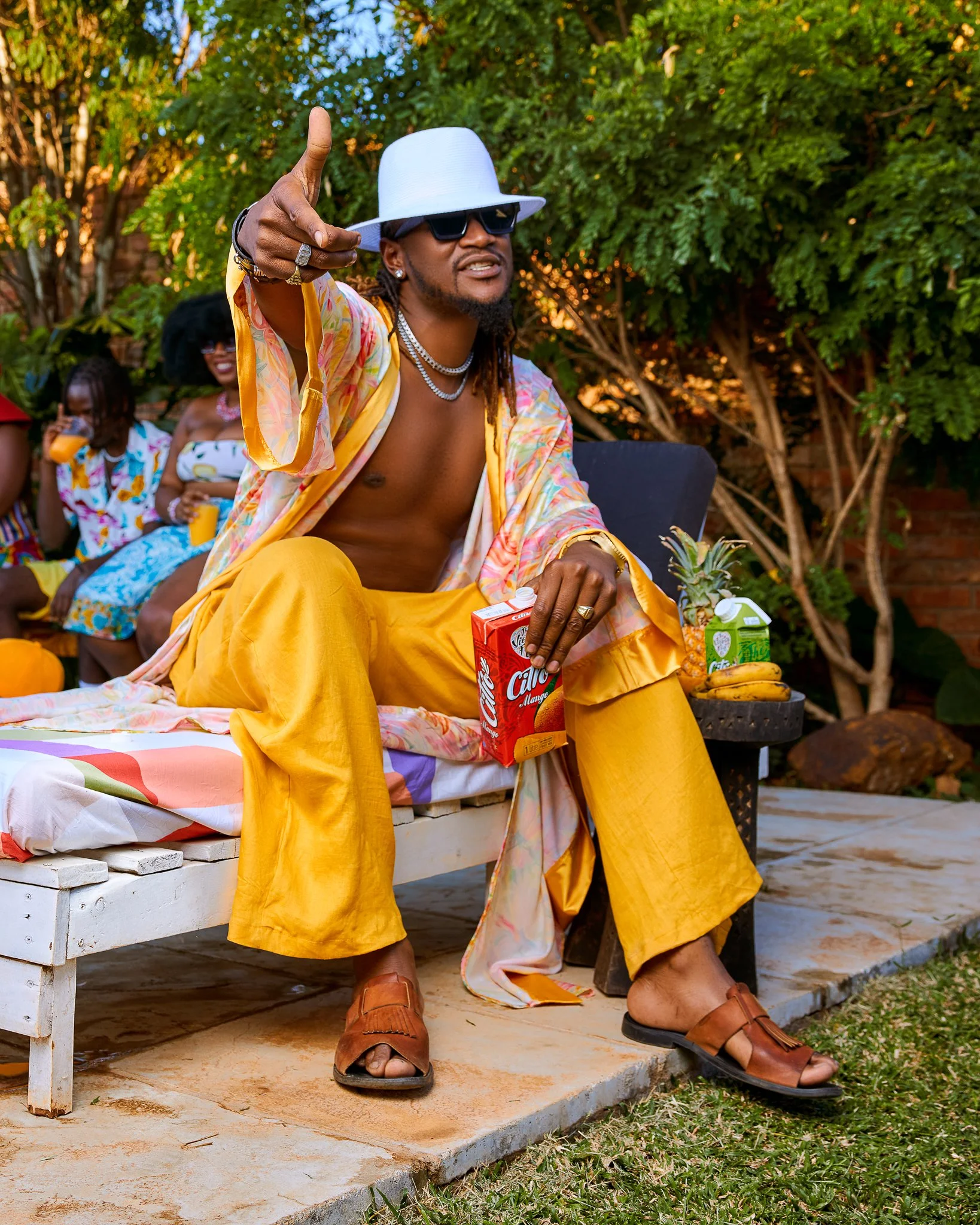 A shirtless man in yellow pants, a floral cover-up, a white hat, and sunglasses sitting on a bench outdoors, holding a juice box and pointing with his right hand.