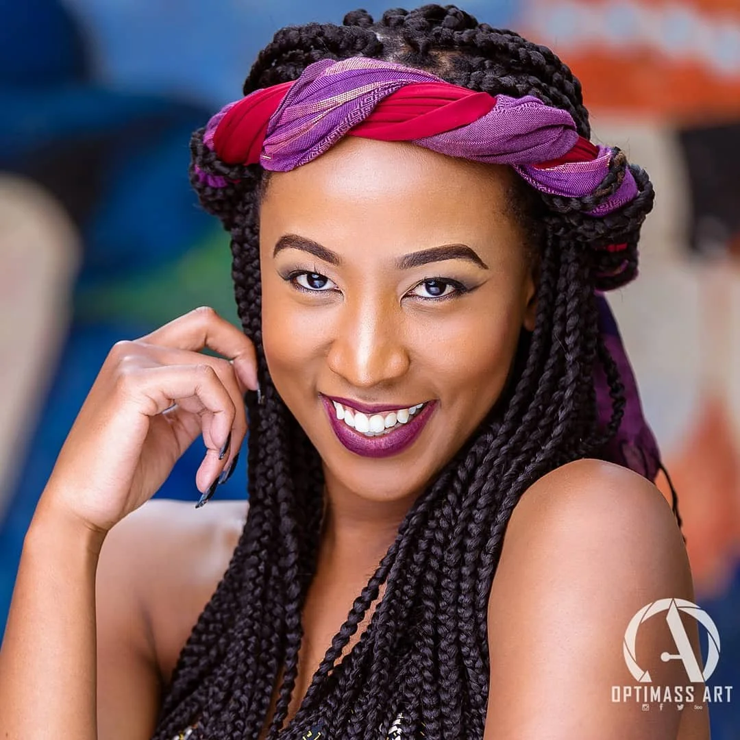 A woman with braided hair wearing a colorful headscarf, smiling and looking at the camera.