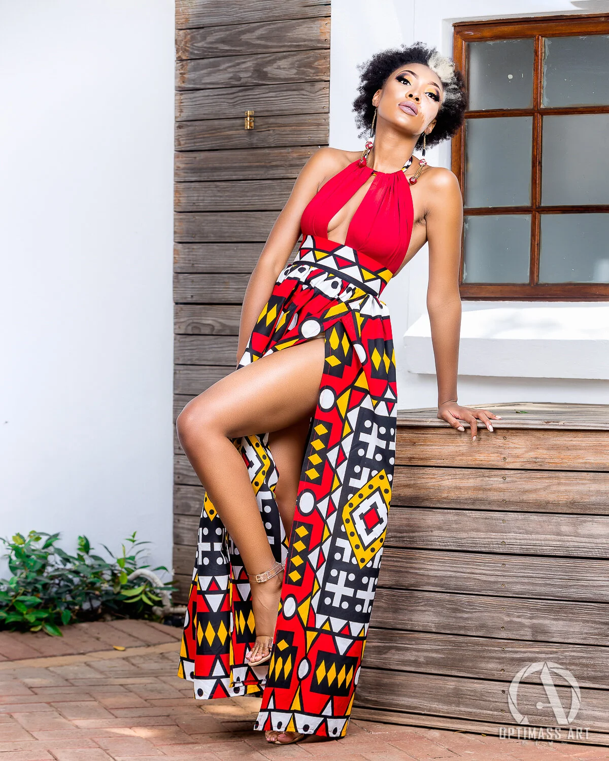 Woman wearing a colorful, patterned high-slit dress standing by a wooden fence, with a window behind her.
