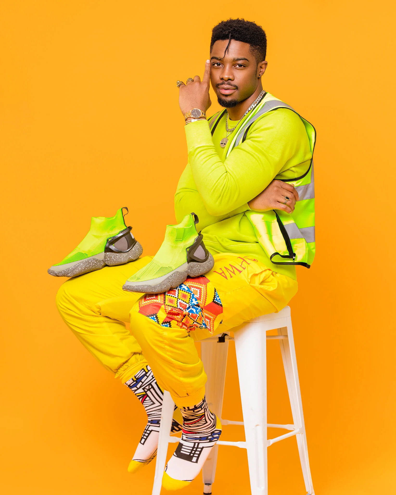 A man sitting on a white stool against a yellow background, wearing neon green and yellow fashion accessories, yellow pants with colorful pattern, and multicolored socks. He has two pairs of bright yellow sneakers with gray soles on his lap and is lo