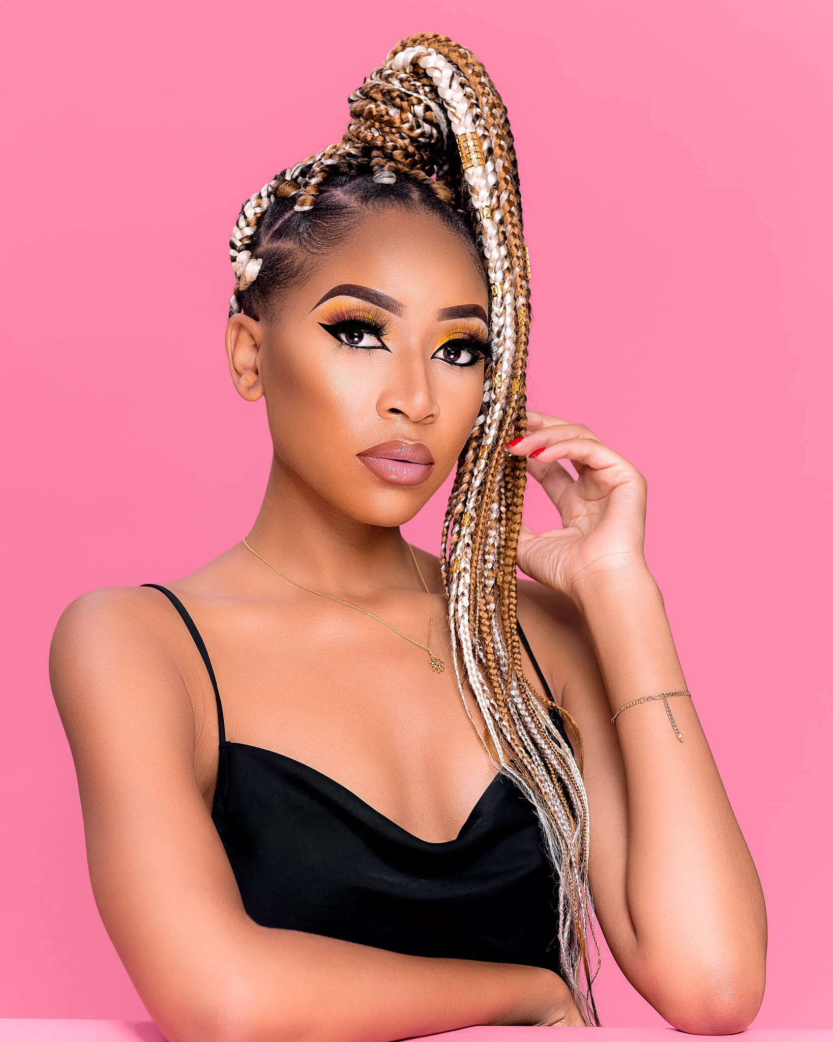 A woman with long blonde and brown braided hair styled in a high braid, wearing a black spaghetti strap top, gold jewelry, and makeup with dramatic eye makeup, posing against a pink background.