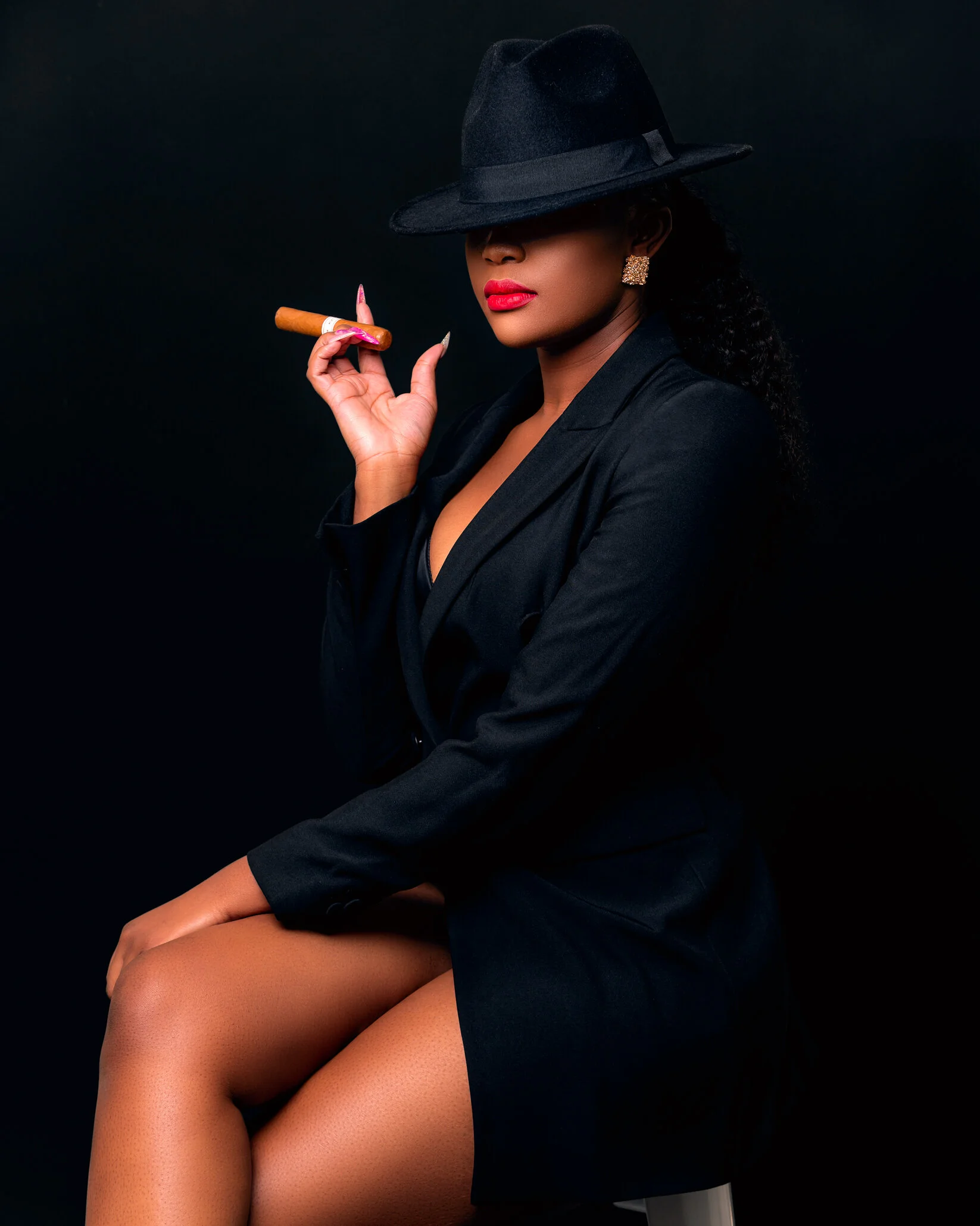 A woman wearing a black coat and hat, holding a cigar, with bright lipstick and earrings, sitting against a black background.