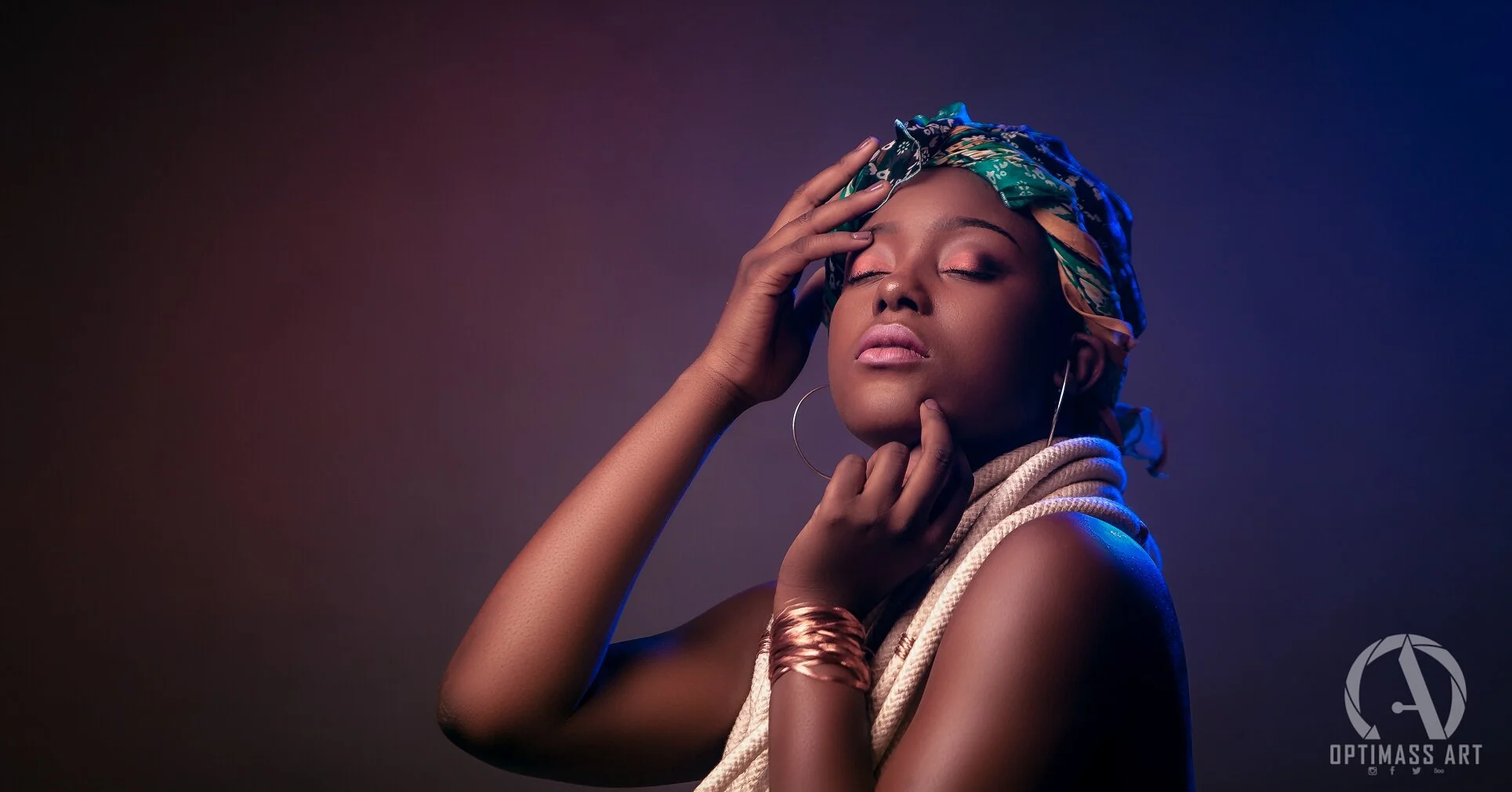 Portrait of a woman with closed eyes, wearing a headscarf, hoop earrings, and jewelry, posing with one hand touching her forehead and the other on her chin against a dark background.