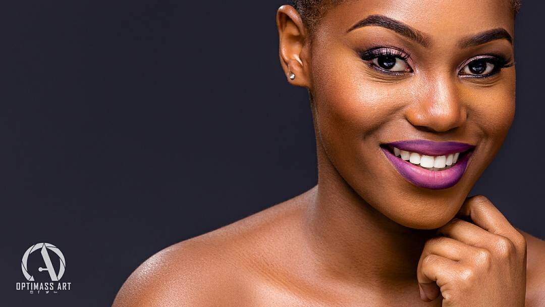 Close-up portrait of a smiling woman with dark hair and purple lipstick, touching her chin with her hand, against a dark background.