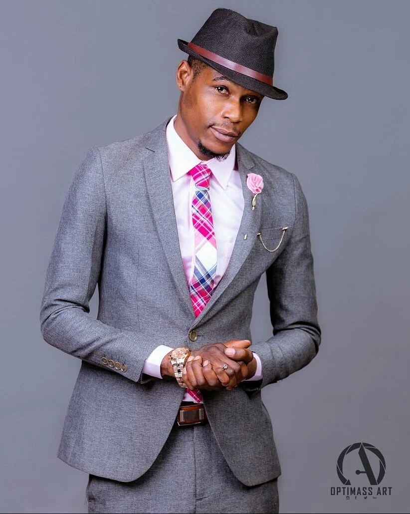 A young man in a gray suit with a pink, red, and white striped tie, wearing a black fedora with a brown band, posing against a plain gray background.