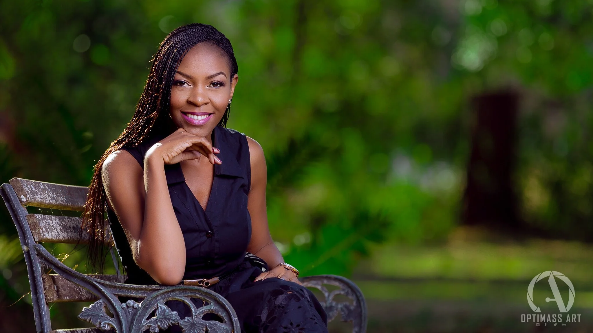 A woman sitting on a weathered metal park bench outdoors, smiling, wearing a sleeveless dark top and pants, with braided hair, in a green, blurred background of trees and foliage.