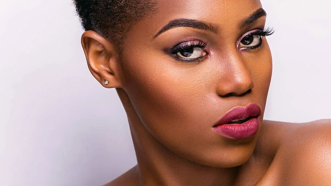 Close-up portrait of a woman with short curly hair, wearing pink eyeshadow and dark lipstick, with small earrings.