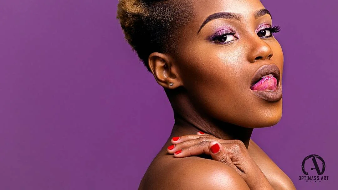 Close-up of a woman with short hair, purple eye makeup, pink lipstick, and red nail polish, posing against a purple background.