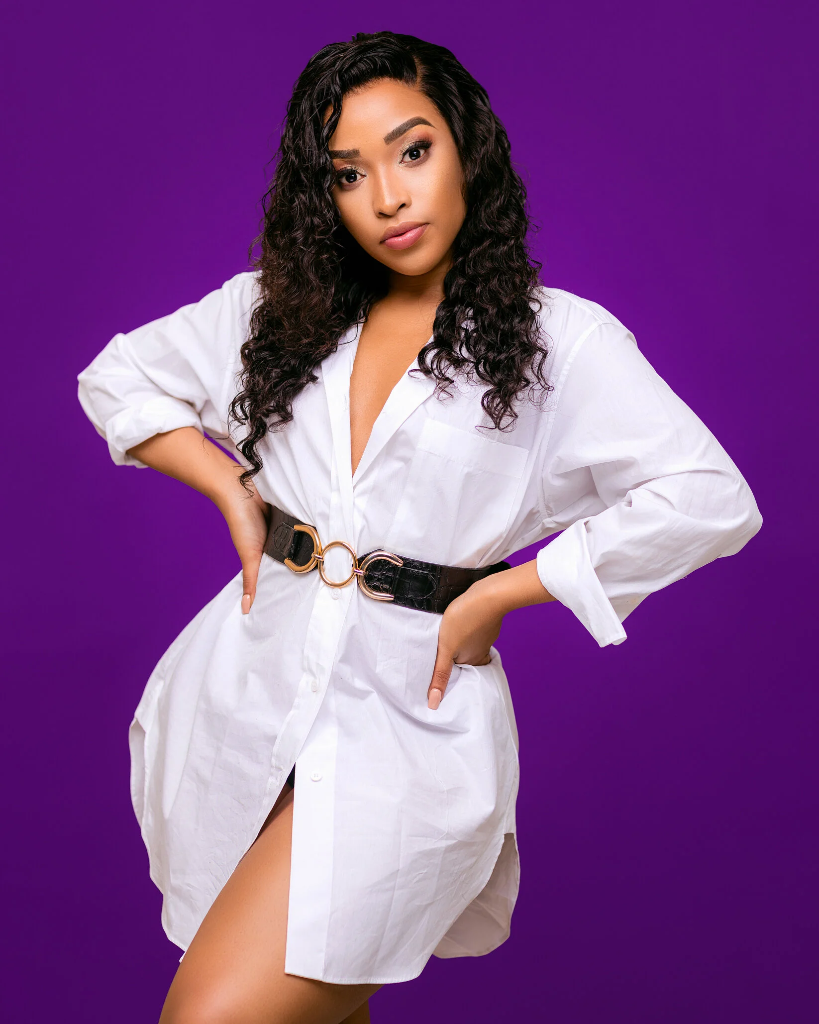 A woman with curly black hair wearing a white button-up shirt dress with a black belt, standing against a purple background.