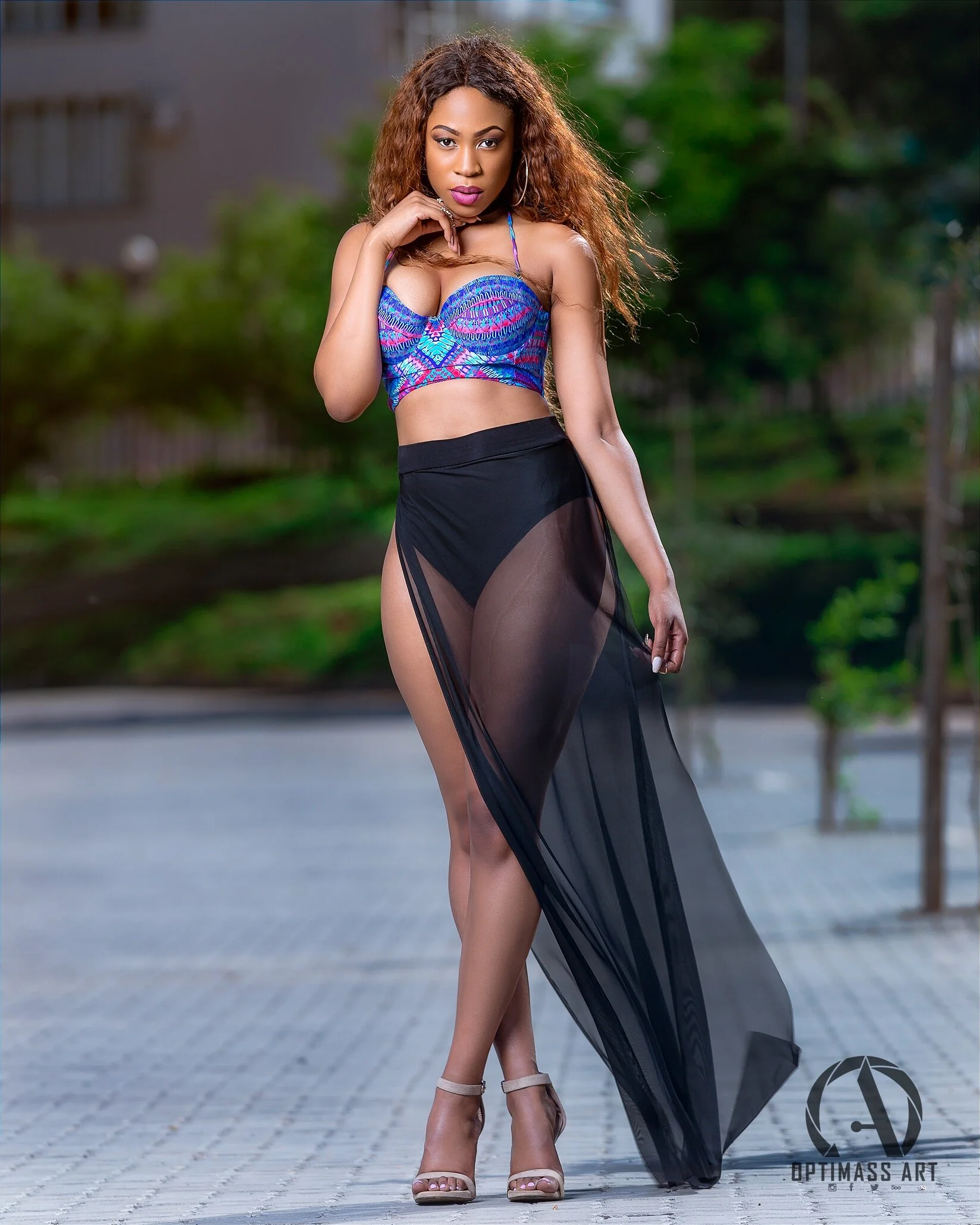 A woman in a colorful crop top and black sheer skirt posing outdoors on a paved surface with trees and buildings in the background.