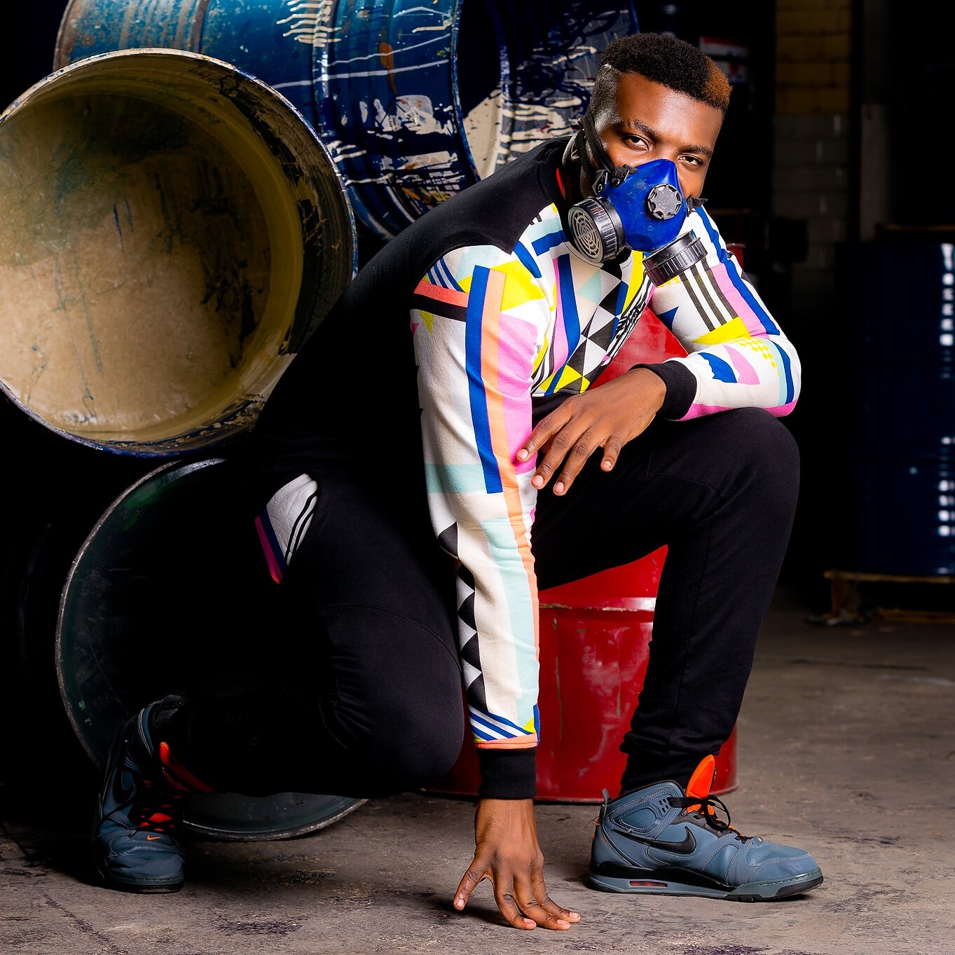 A man wearing a colorful geometric-patterned jacket, black pants, and sneakers crouches on the ground with one hand touching the floor while wearing a blue respirator mask, with large industrial drums in the background.