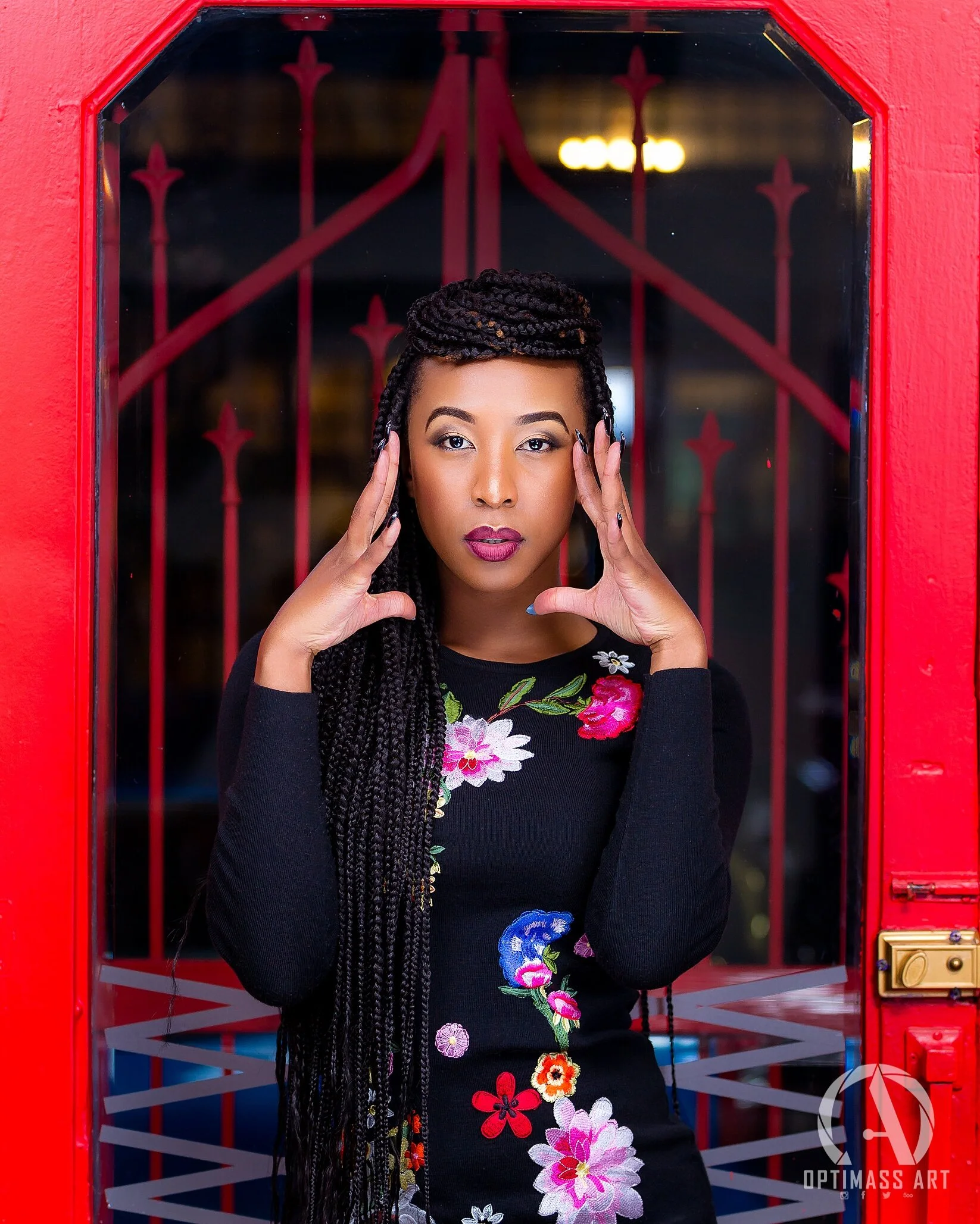 A woman with long braided hair standing in front of a red door with a starburst pattern, wearing a black floral dress and striking a pose with her hands on the sides of her head.